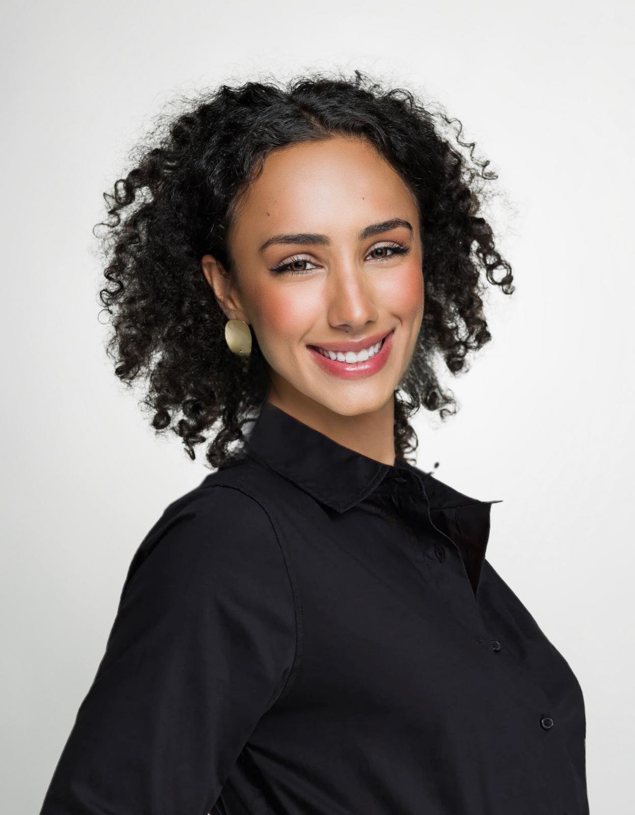 A woman smiling with curly black hair, wearing a black button-up shirt and cream-colored earrings, against a plain light background.