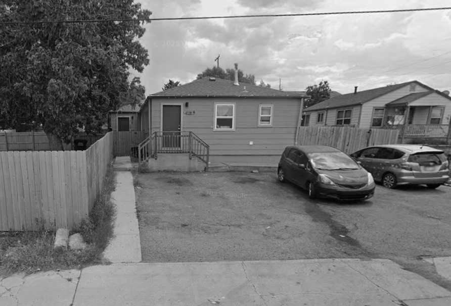 A small, single-story house with green siding and a gray roof. There's a wooden ramp and steps leading to the front door, with a wooden fence on either side. Two cars are parked in front of the house on a dirt parking area. The sky is cloudy, and neighboring houses are visible in the background.