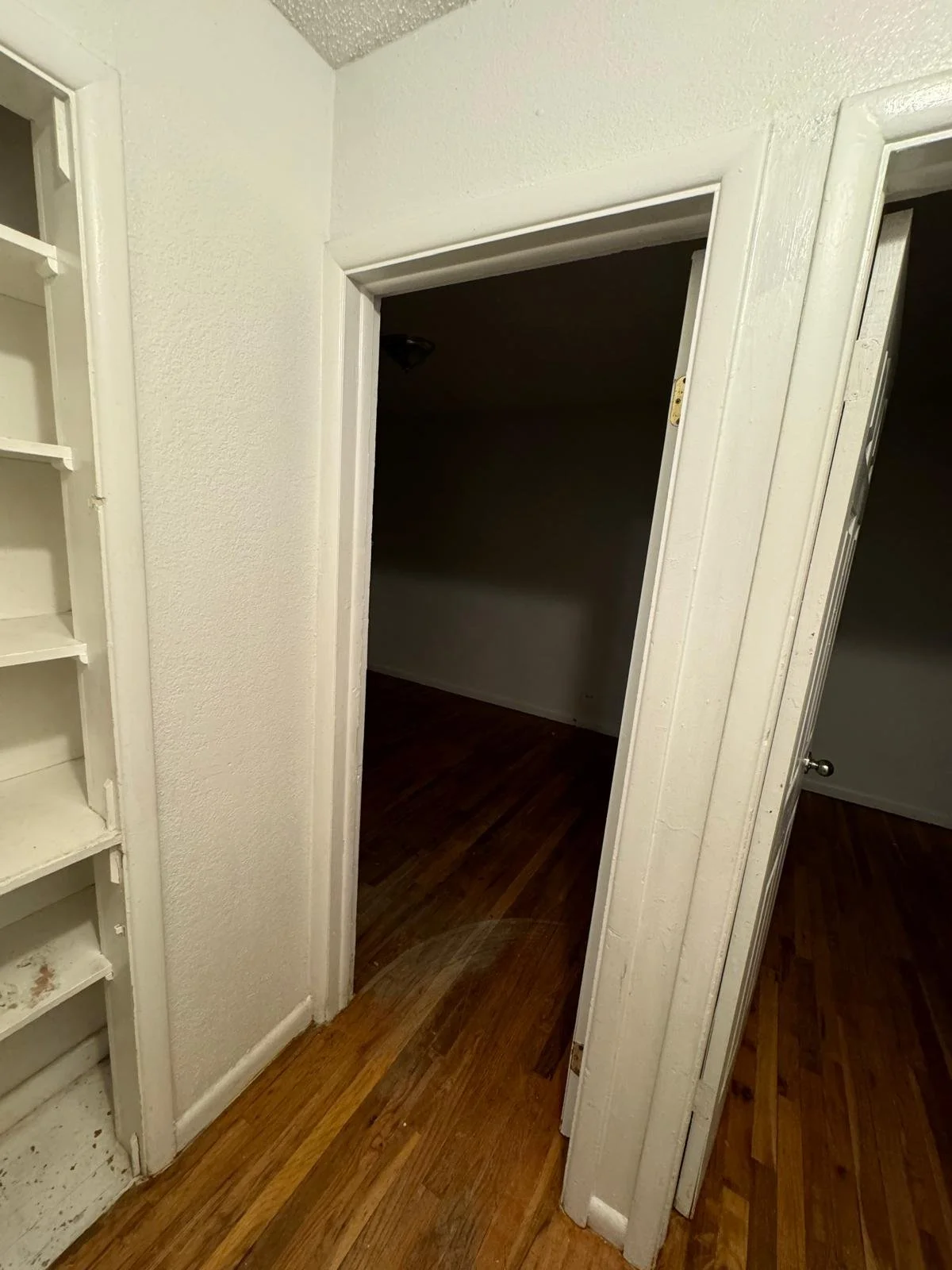 Interior view of an empty room with hardwood floors, white walls, and a doorway leading into a darker room or closet.