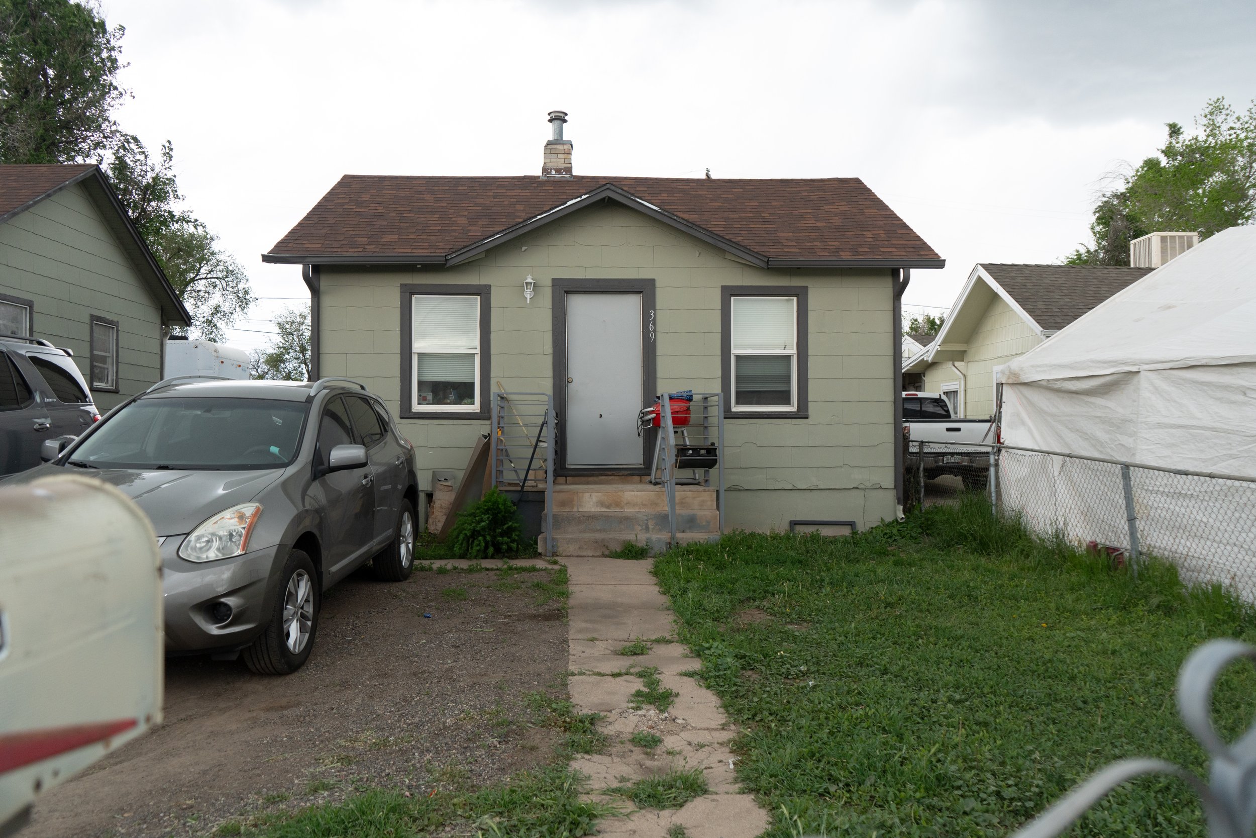 A small, green, single-story house with a brown shingled roof. There are two windows on the front, a door with the number 369, and a short set of steps leading to the entrance. A gray car is parked on the driveway to the left of the house, and there 