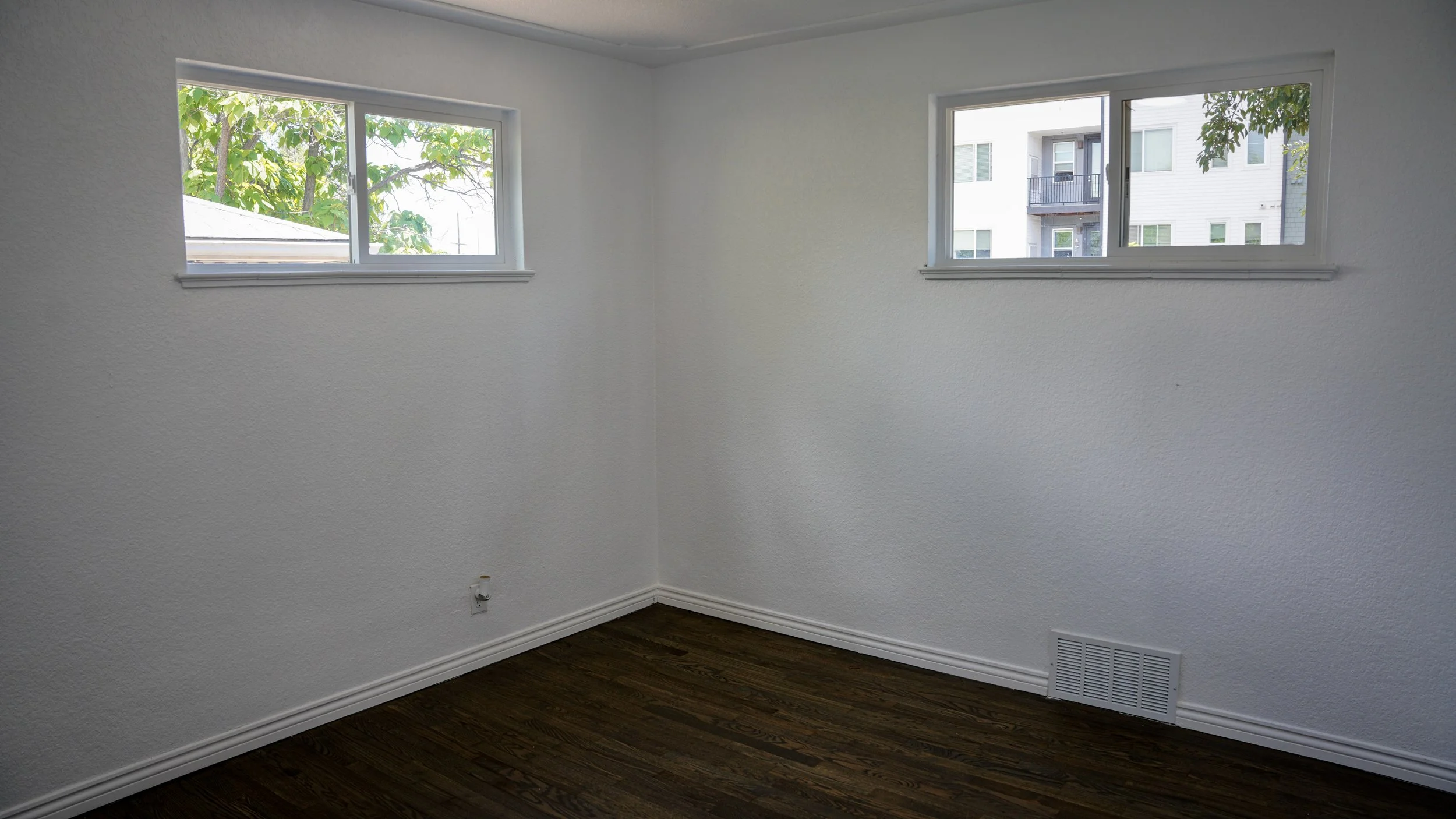 Empty room with white textured walls, dark wood flooring, two side-by-side rectangular windows with white frames, and an air vent near the floor on one wall.