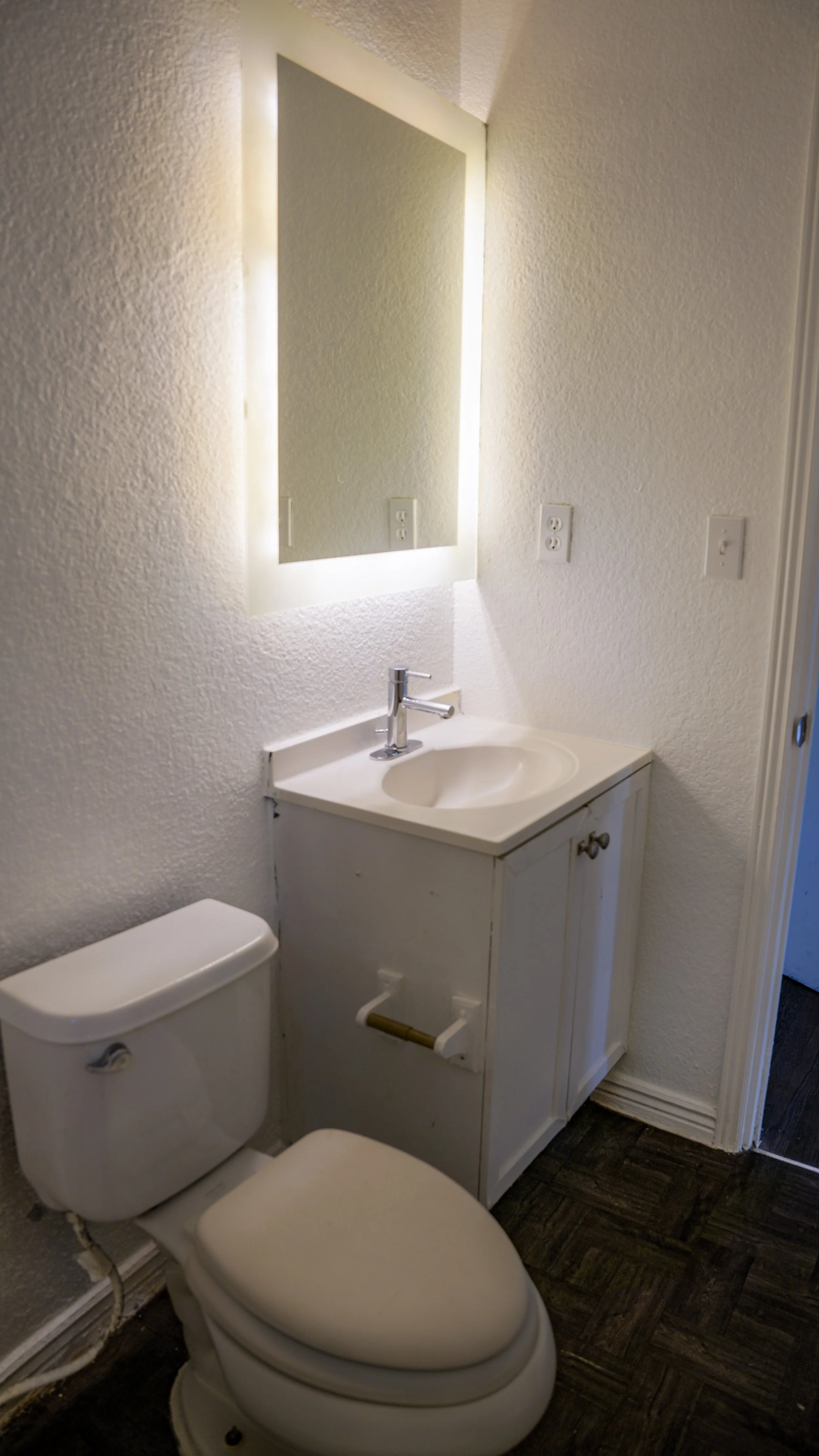 A small bathroom with a white toilet, a white sink with a cabinet, and a rectangular mirror with backlit lighting. There are three electrical outlets on the wall, and the floor is dark wood or laminate.
