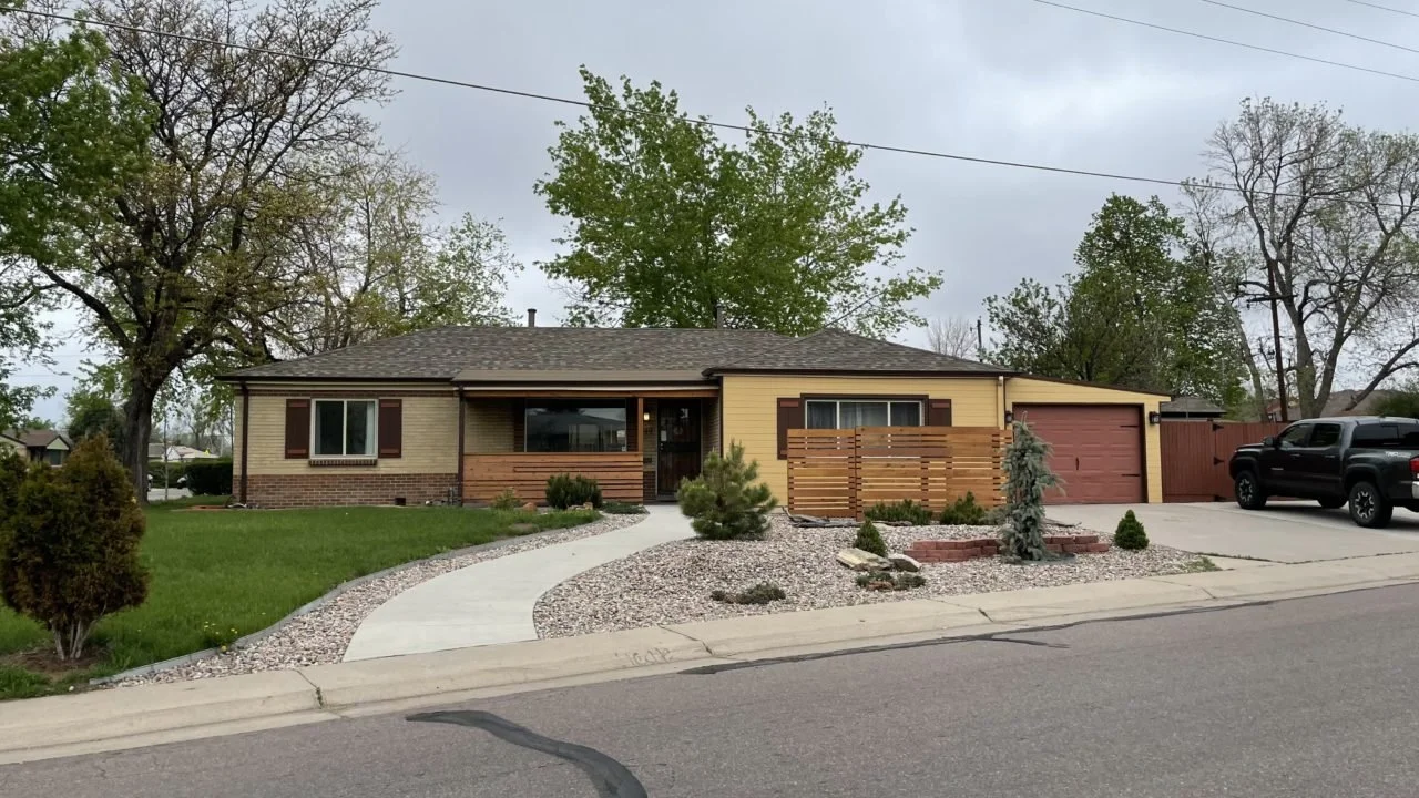 Front view of a single-story house with a curved walkway, green lawn, and landscaped yard with small shrubs and trees, and a black truck parked in the driveway.