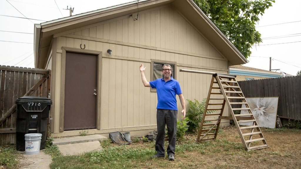 Man in blue shirt standing in backyard, smiling, waving, next to a small beige shed with a ladder leaning against it, surrounded by a wooden fence and some plants.