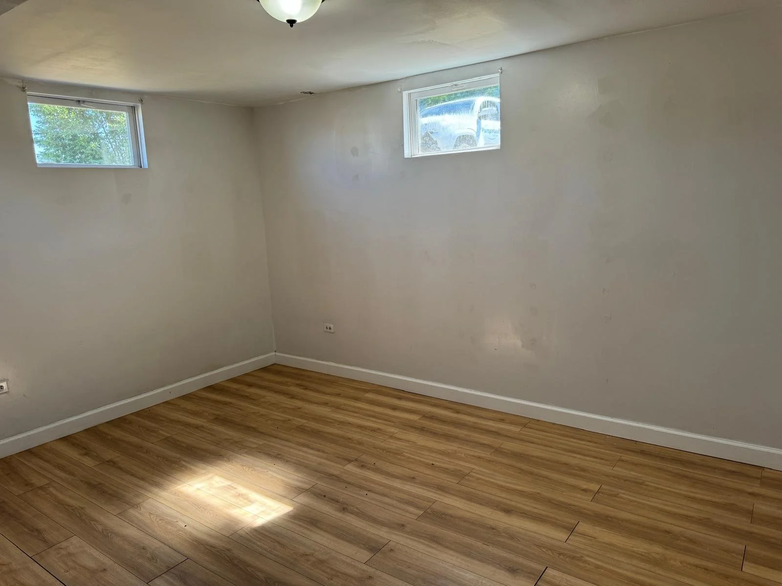 Empty room with light-colored walls, hardwood flooring, two small horizontal windows, and a ceiling light fixture.