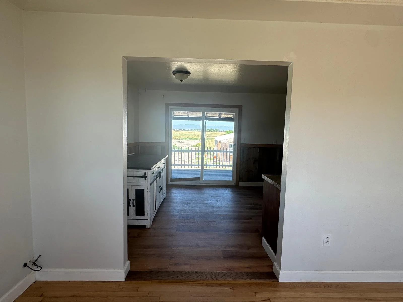Empty room with white walls and hardwood floors leading to a kitchen space with a view of a deck and landscape through sliding glass doors.
