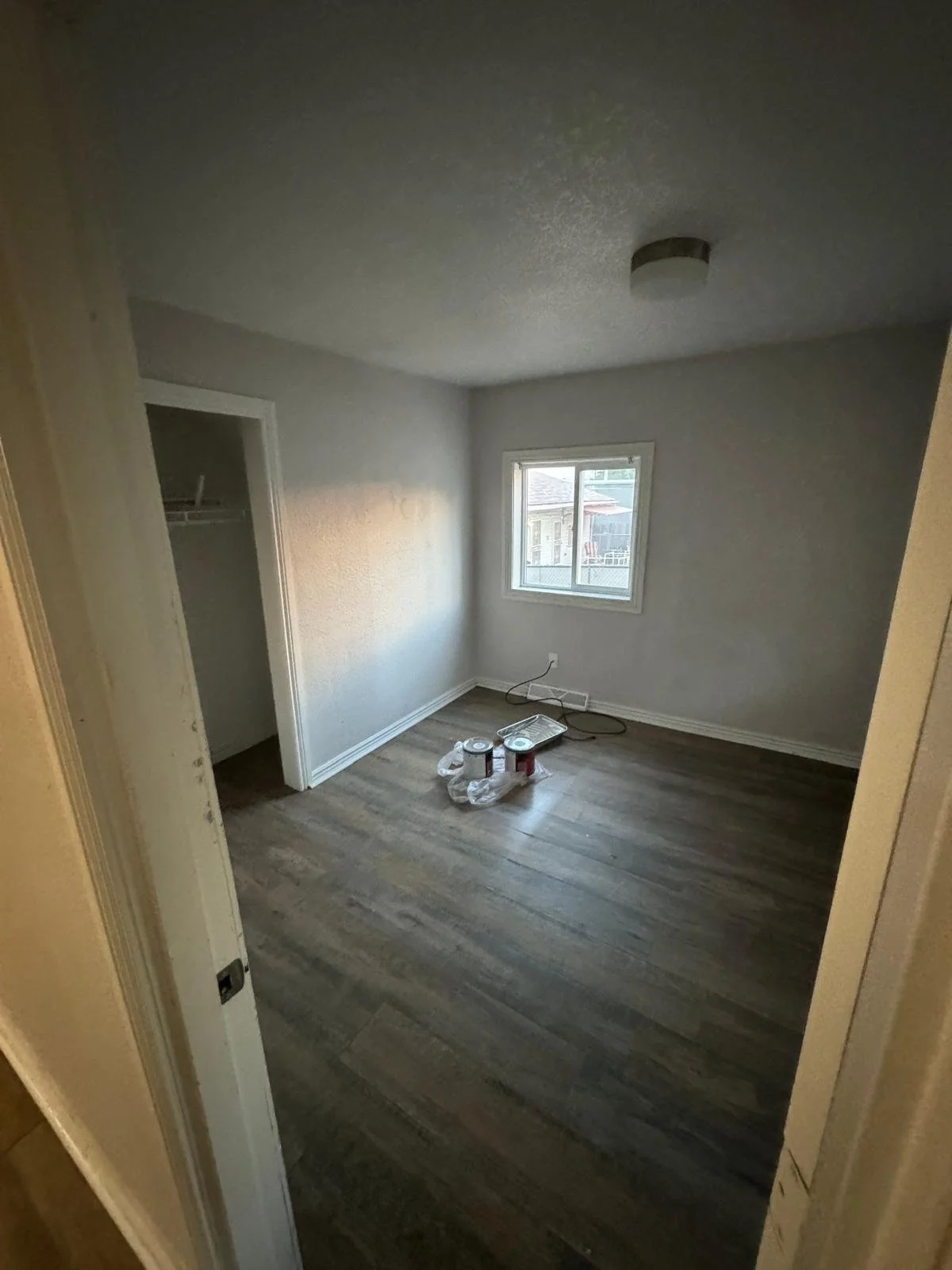 Empty room with hardwood flooring, white walls, a window, a closet, and paint supplies on the floor.