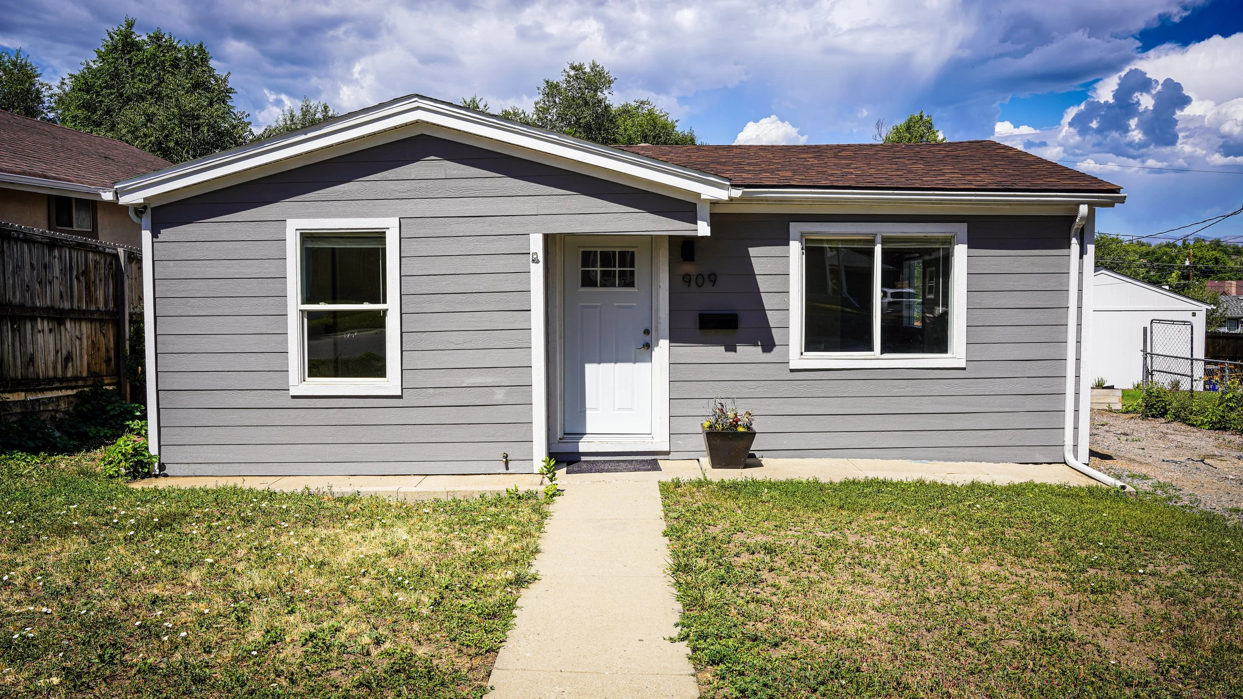 Front view of a gray house with white trim, two windows, a white door, and a small pathway leading to the entrance, with a lawn and a neighboring house in the background.