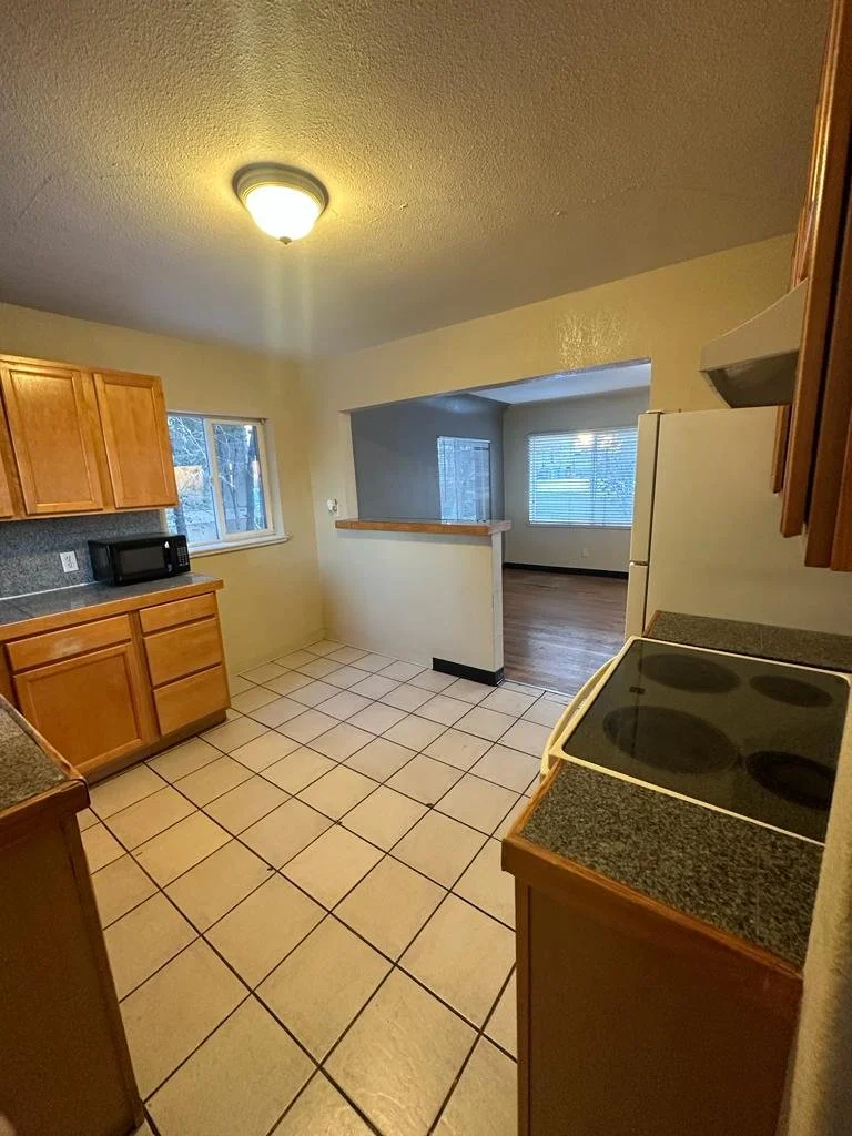 Kitchen with wooden cabinets, tiled floor, and a white refrigerator. A window above the counter, a microwave on the counter, and a stove with four burners. Open view into an adjacent room with hardwood flooring and windows.