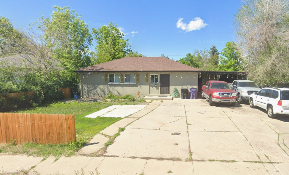 A single-story house with a brown roof and gray walls, with a small fenced garden area and a concrete driveway with parked cars. There are trees in the background under a blue sky with a few clouds.
