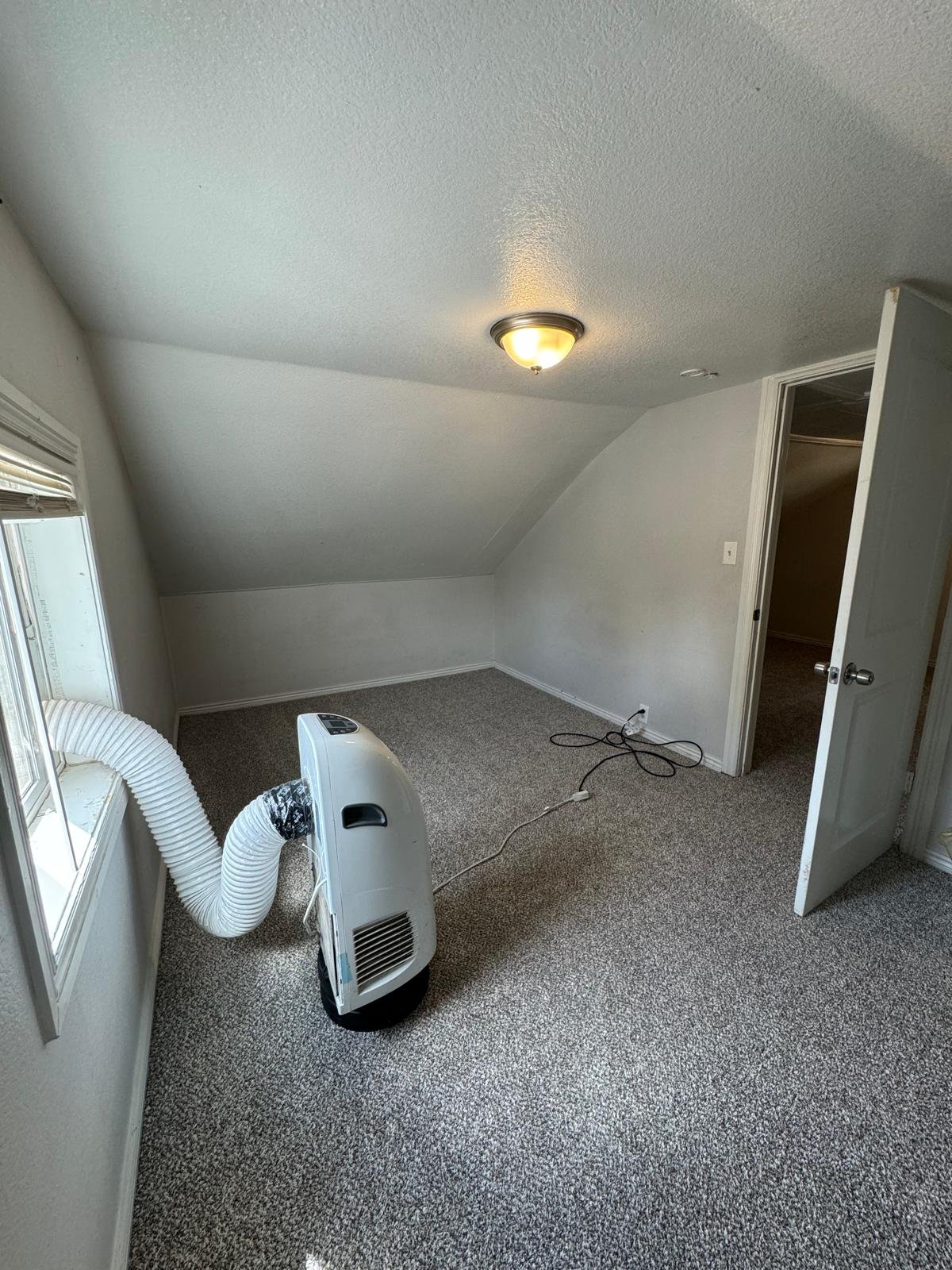 Empty small room with beige carpet, sloped ceiling, a window with blinds, a closed door, and a portable air conditioner with a hose vacuuming air outside.