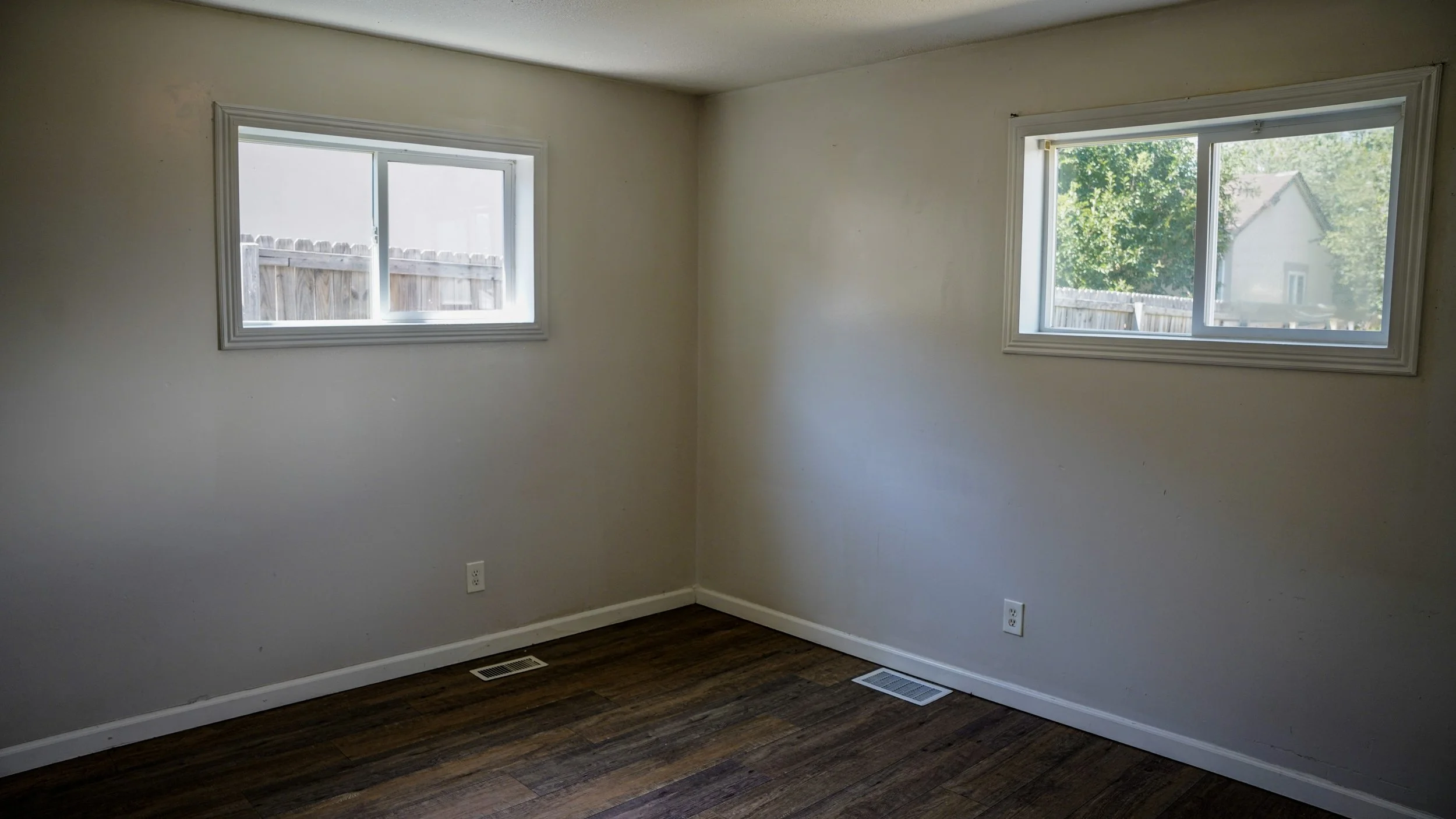 Empty room with white walls, two windows, wood flooring, and a ceiling vent.