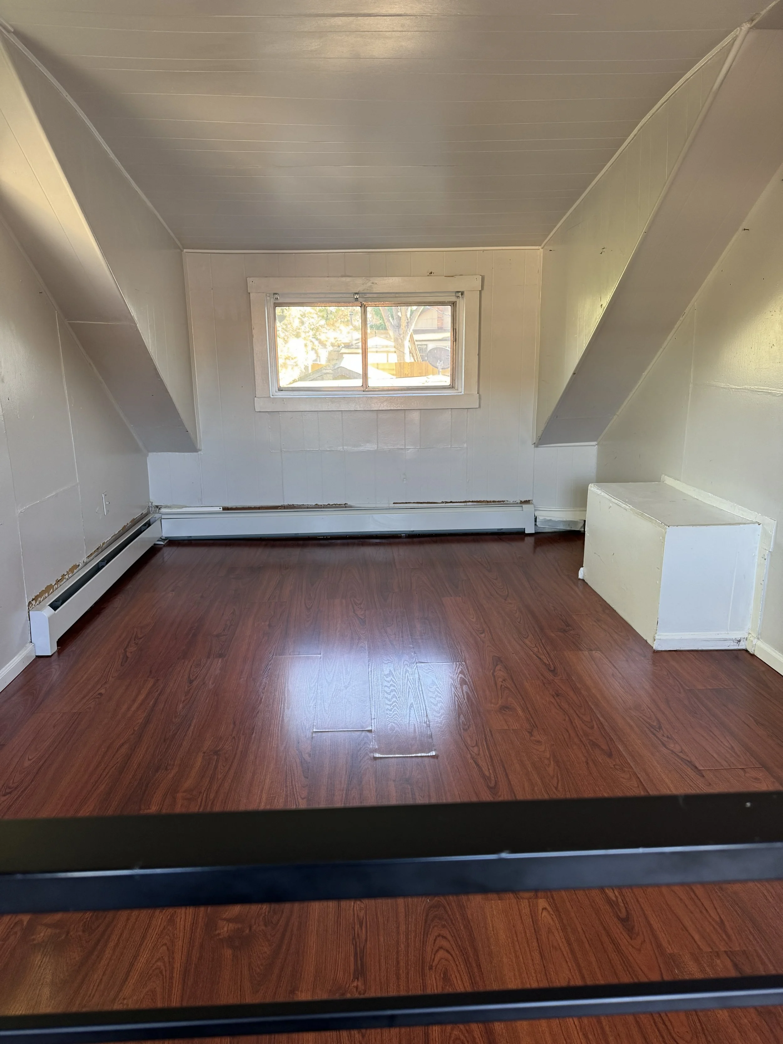 Empty room with white paneled walls, hardwood floor, a small window, and sloped ceiling.