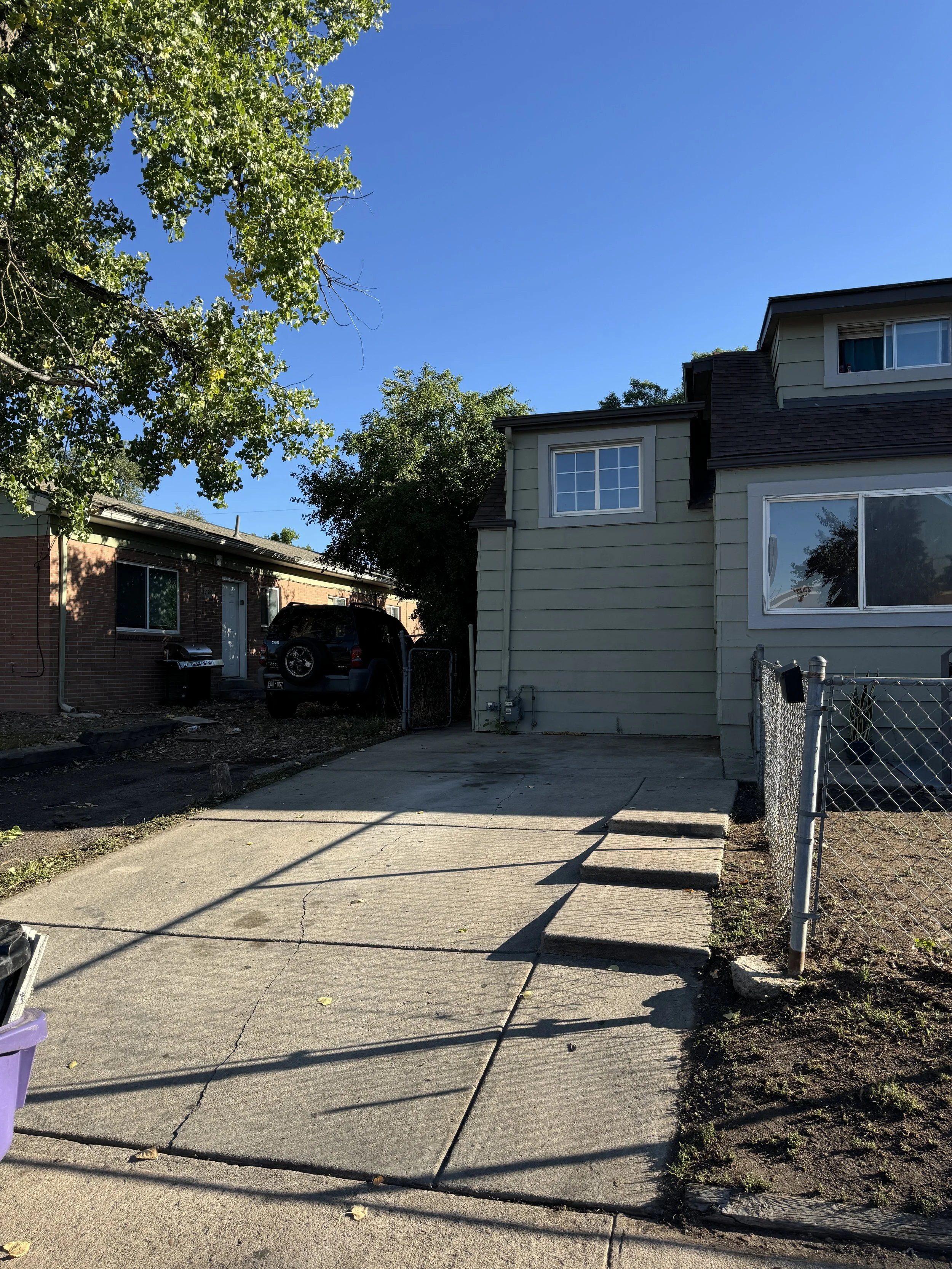 Empty concrete driveway with steps leading to a house, chain-link fence on the right, parked black SUV on the left, house with beige siding and a window, trees and blue sky in the background