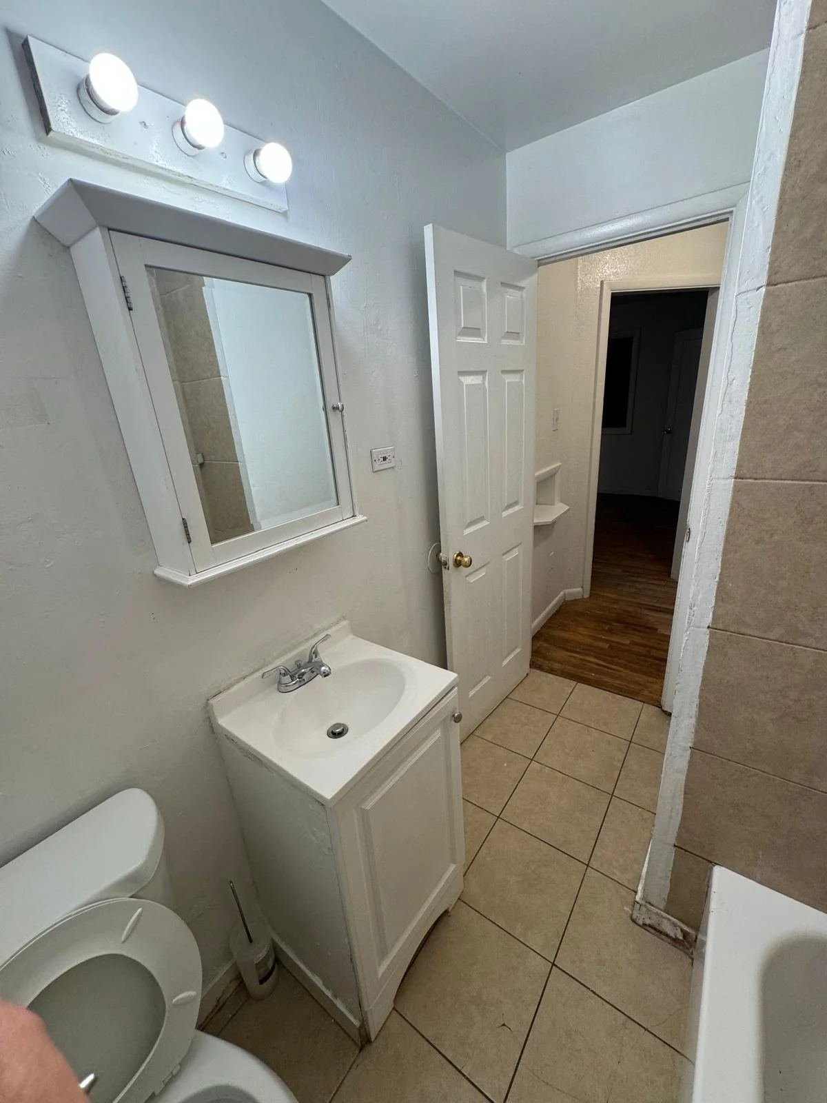 A small bathroom with a toilet, a white vanity with a small sink, a mirror cabinet, a light fixture with three bulbs, and a door leading to a room with wooden flooring.