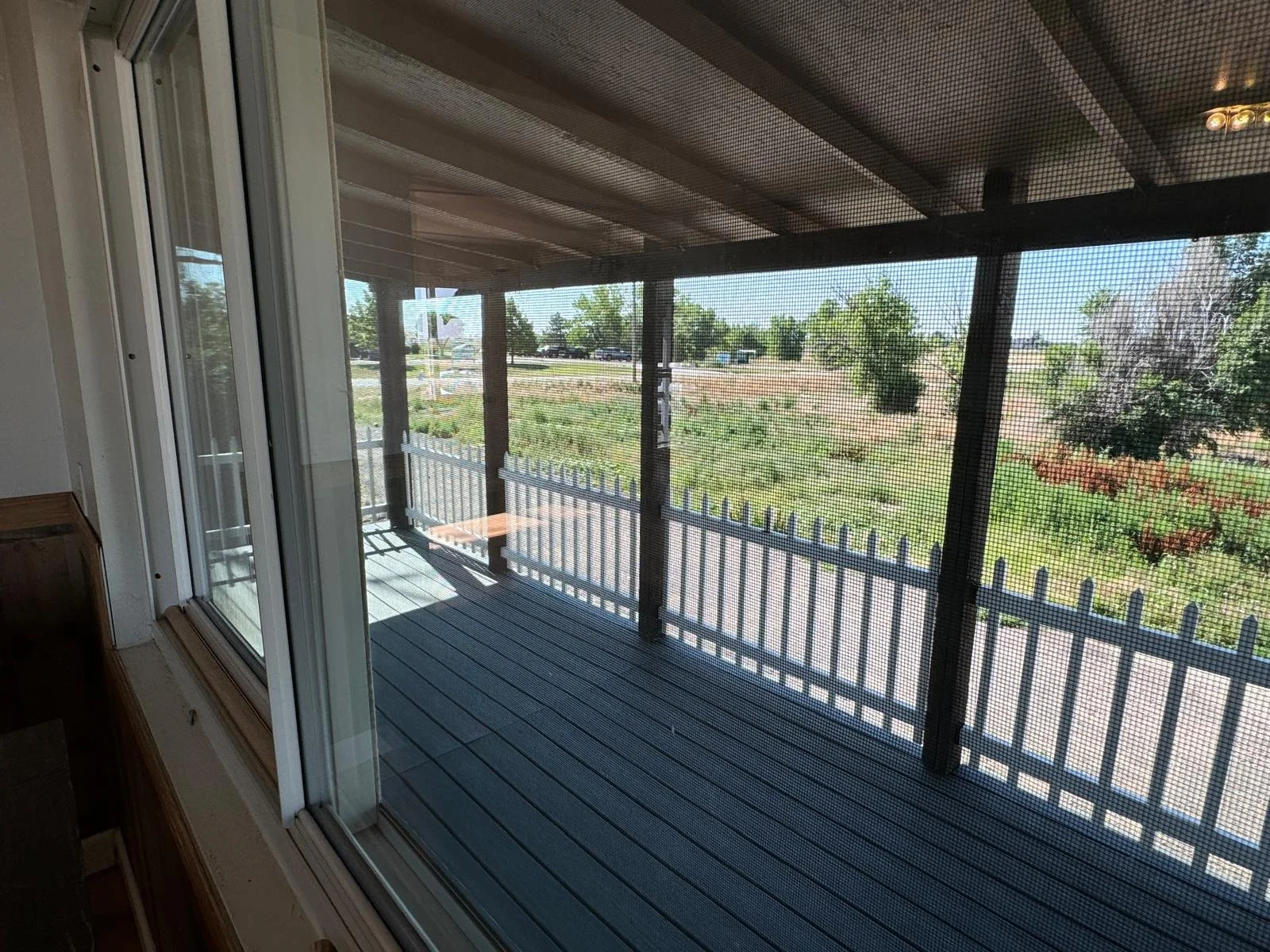 View through a screened-in porch with a wooden deck, metal railing, and trees and open landscape beyond.