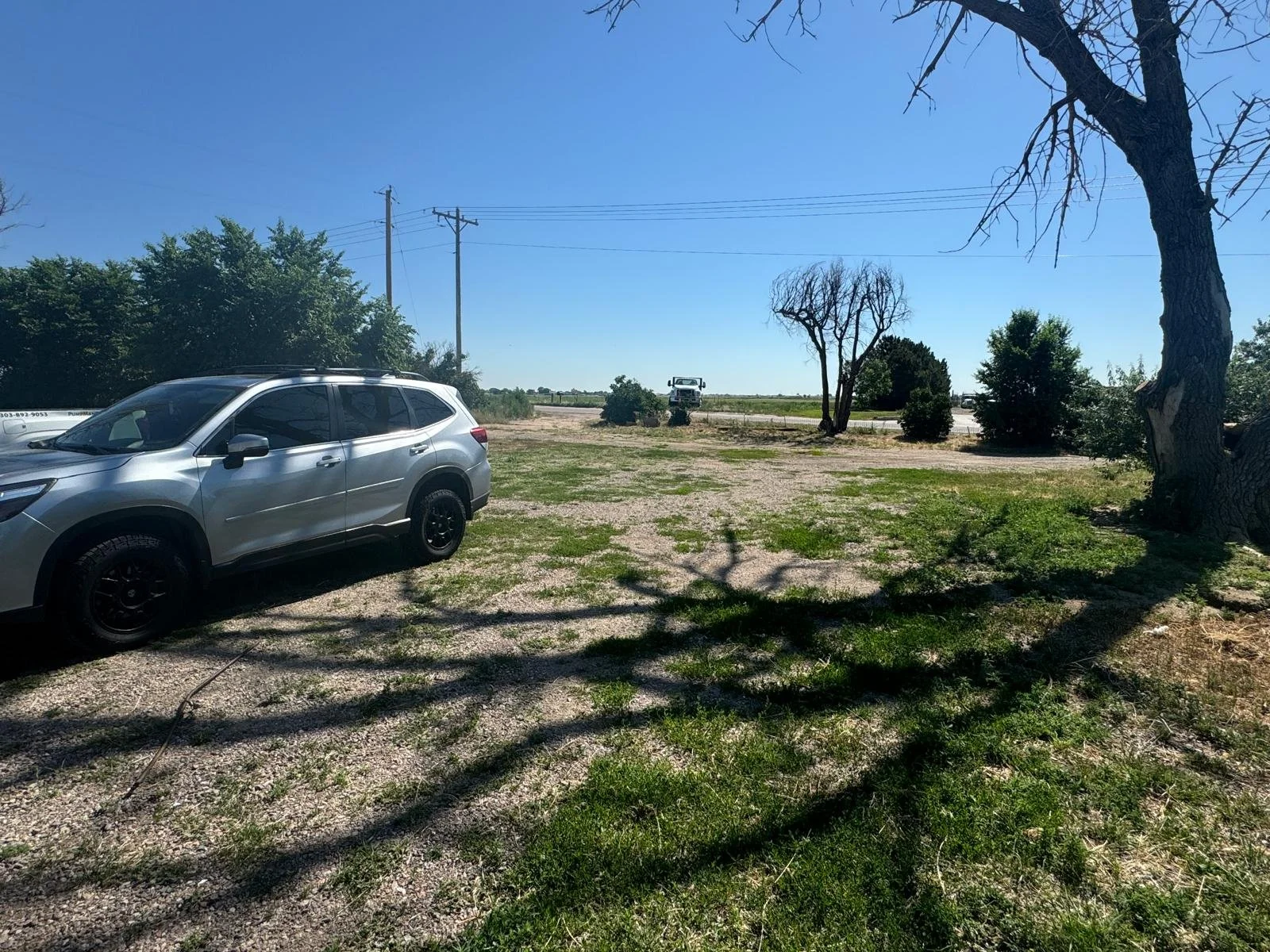 A parking area with a silver SUV parked on gravel, shadows of trees on the ground, a few small trees and shrubs, utility poles with power lines, and open land with a clear blue sky.