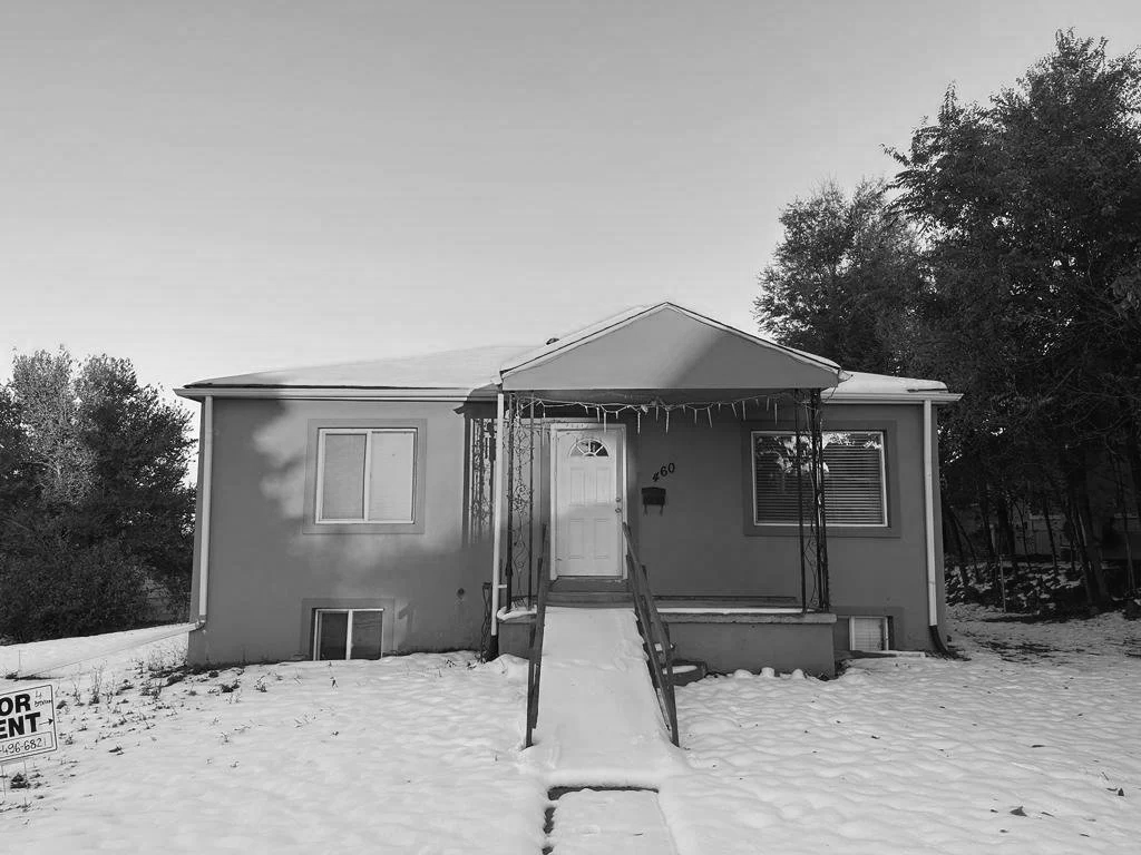 A small house with a covered front porch, a white door, and two windows, surrounded by snow and trees, with a 'For Rent' sign in the yard.