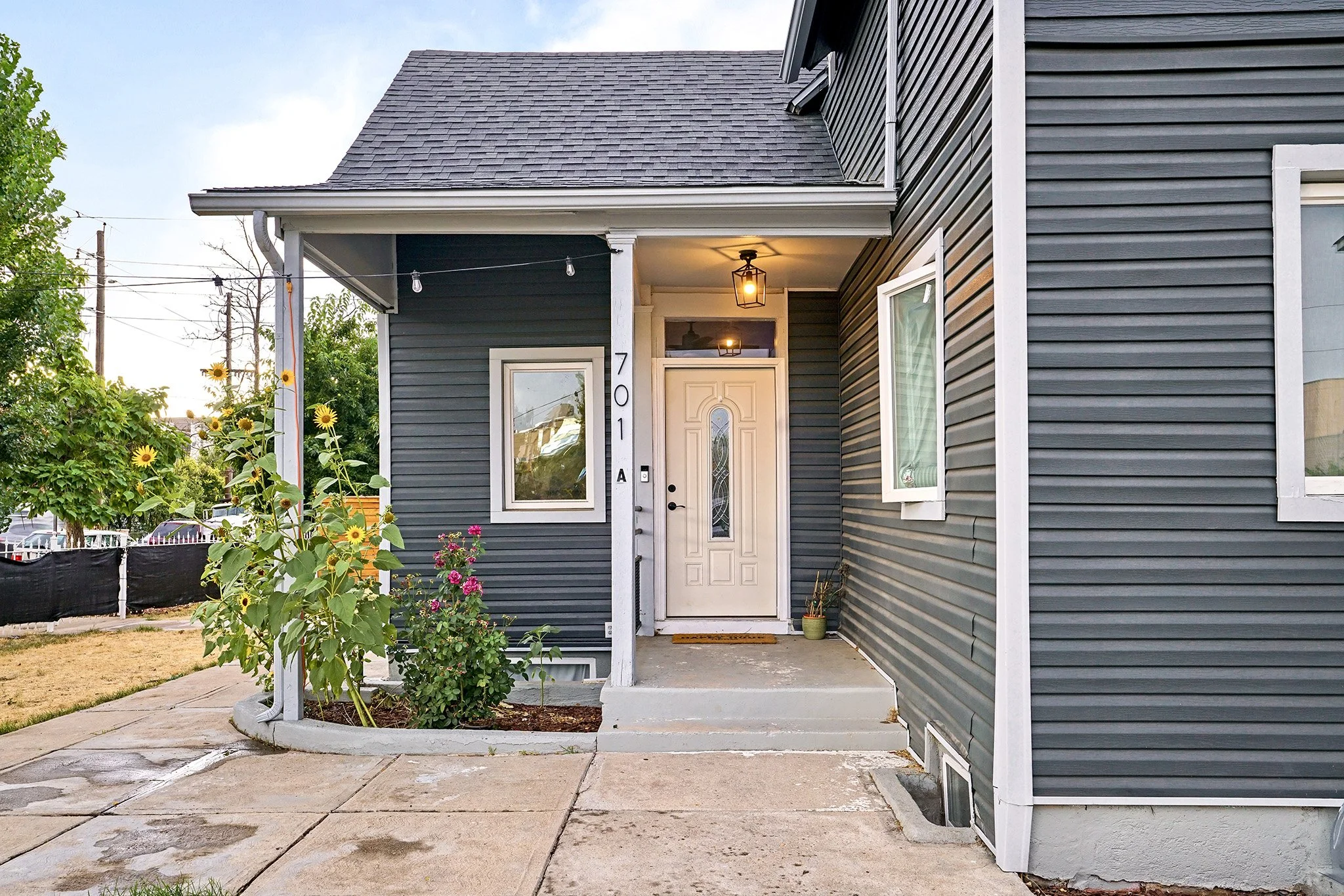 Front porch of a house with dark gray siding, white door with decorative glass, potted plants, and sunflowers, with a concrete walkway and steps leading up to the door.