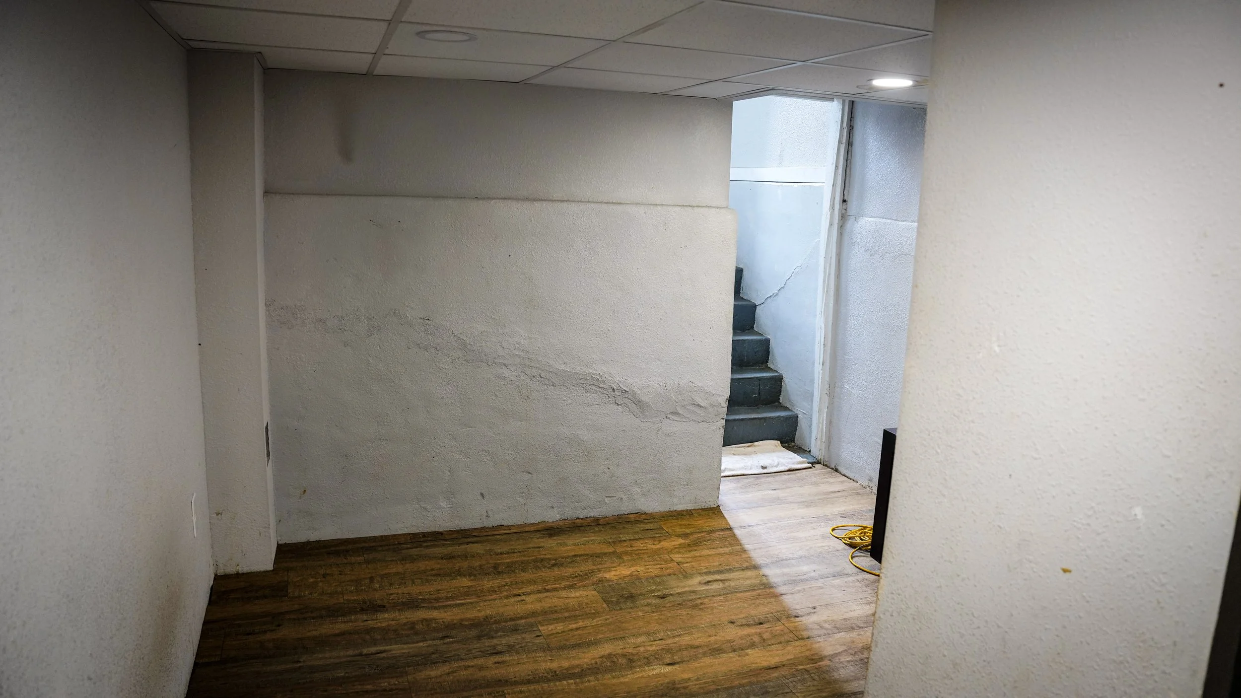 Interior of a basement with white walls, a wooden floor, a set of stairs leading up, and a small crack on the wall.
