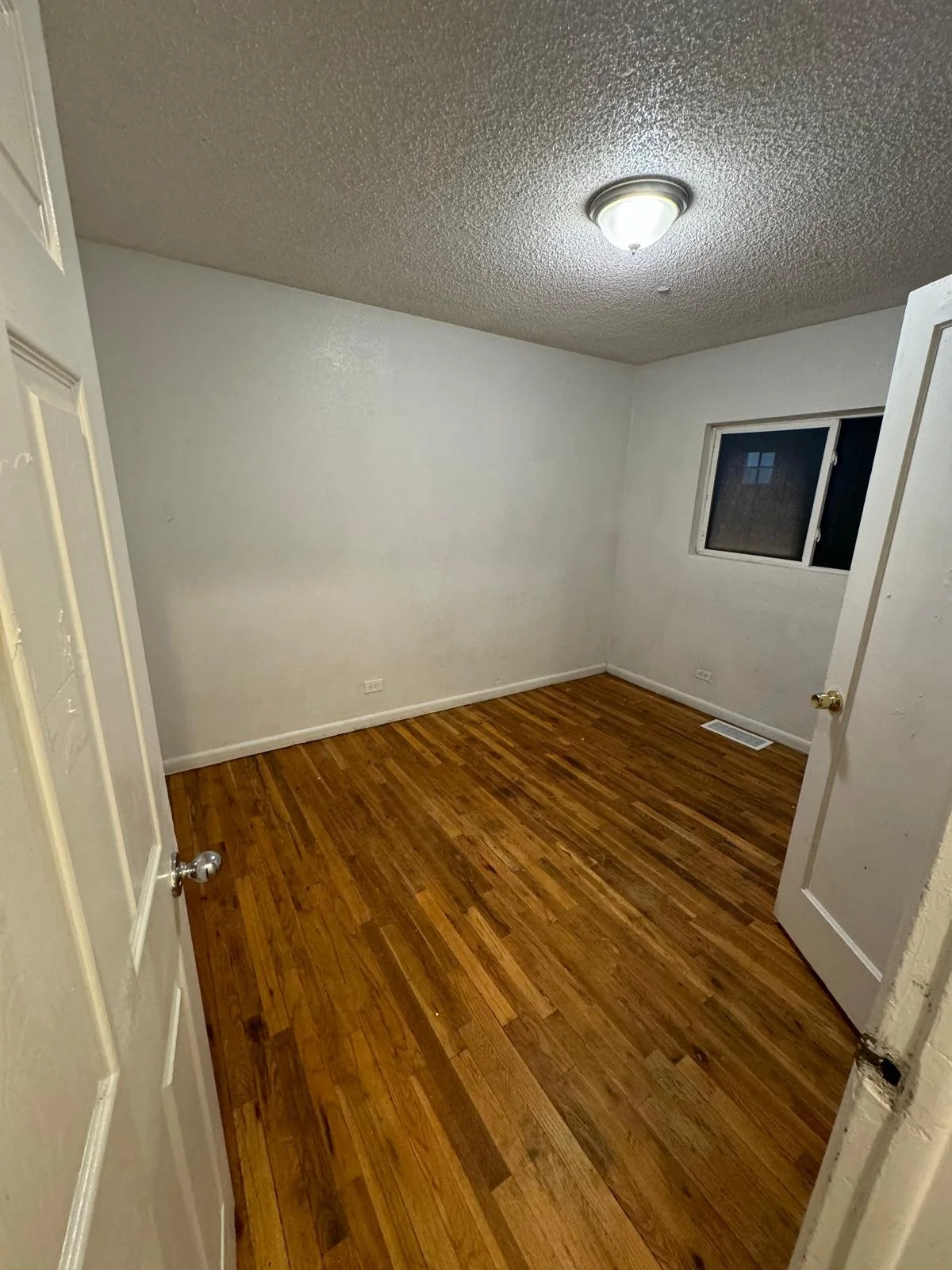 Empty room with hardwood floor, white walls, window, ceiling light, and open door.