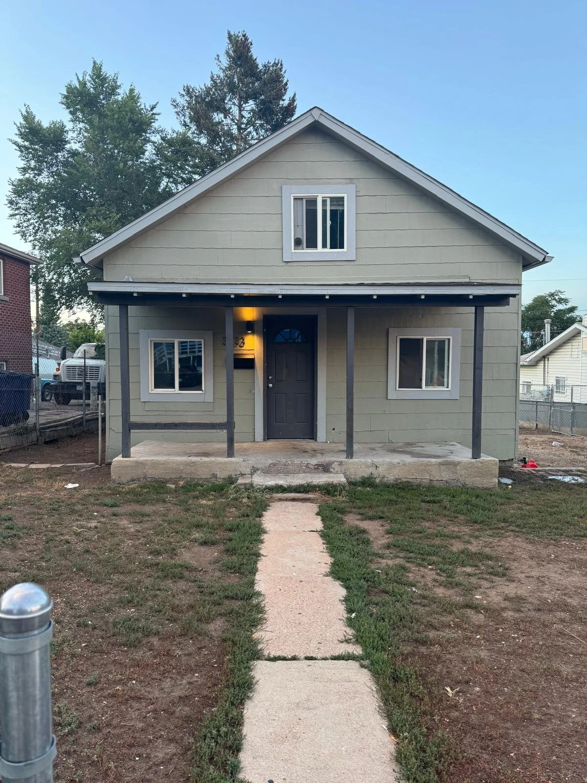 Front of a small, light green house with a dark front door and a porch supported by two dark gray posts. There is a walkway leading to the porch, with patches of grass and dirt on either side. The house has two windows on the front, and an upper wind