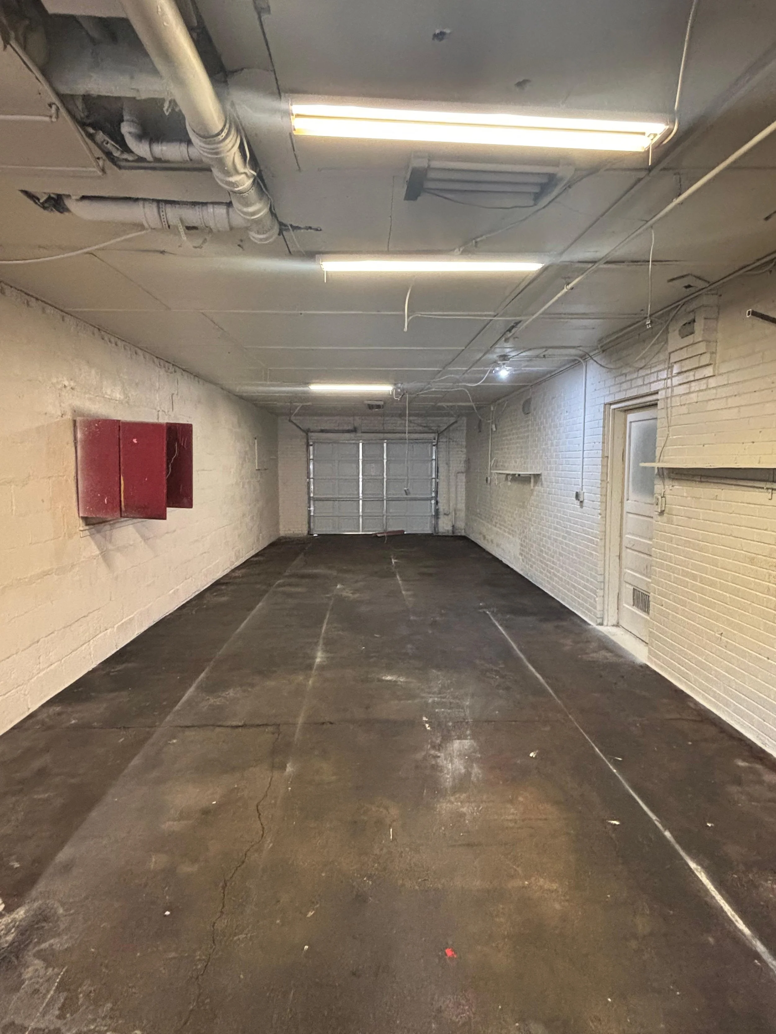 Empty garage with concrete floor, brick walls, a closed garage door at the end, fluorescent lights on the ceiling, and a red metal cabinet on the left wall.