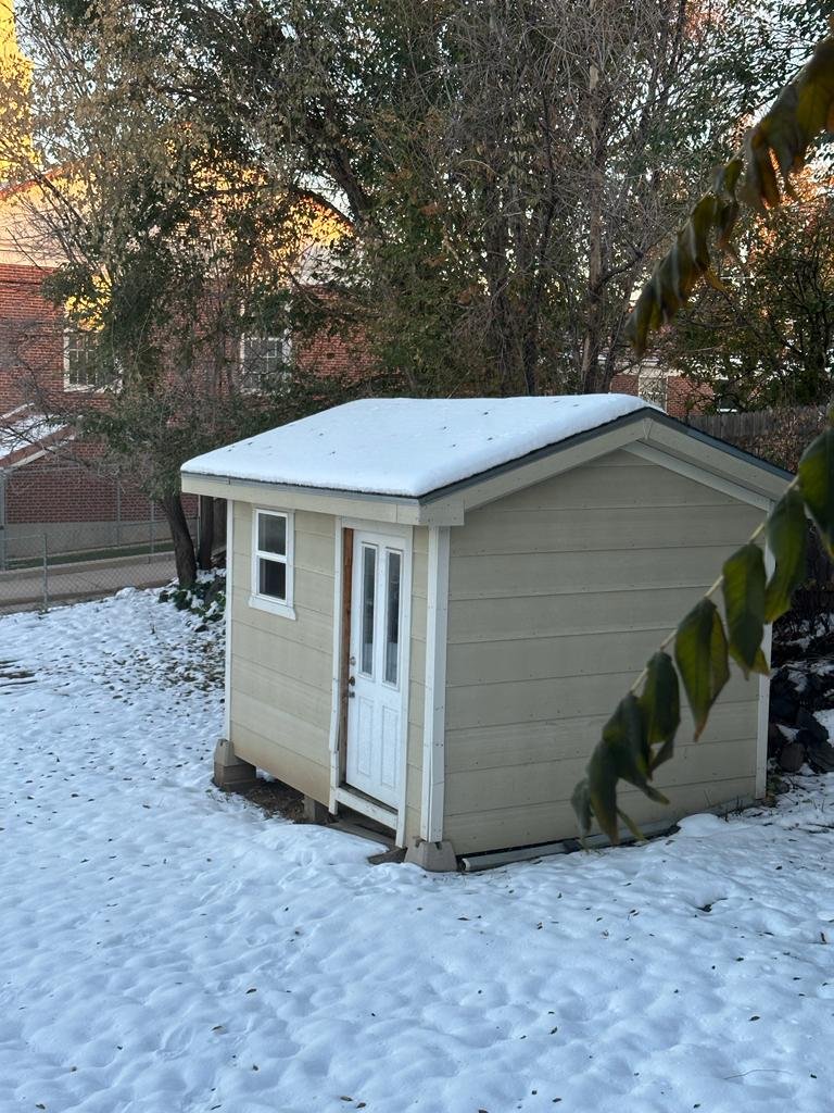 Small shed with a shingled roof covered in snow in a backyard, with trees and a brick building in the background.