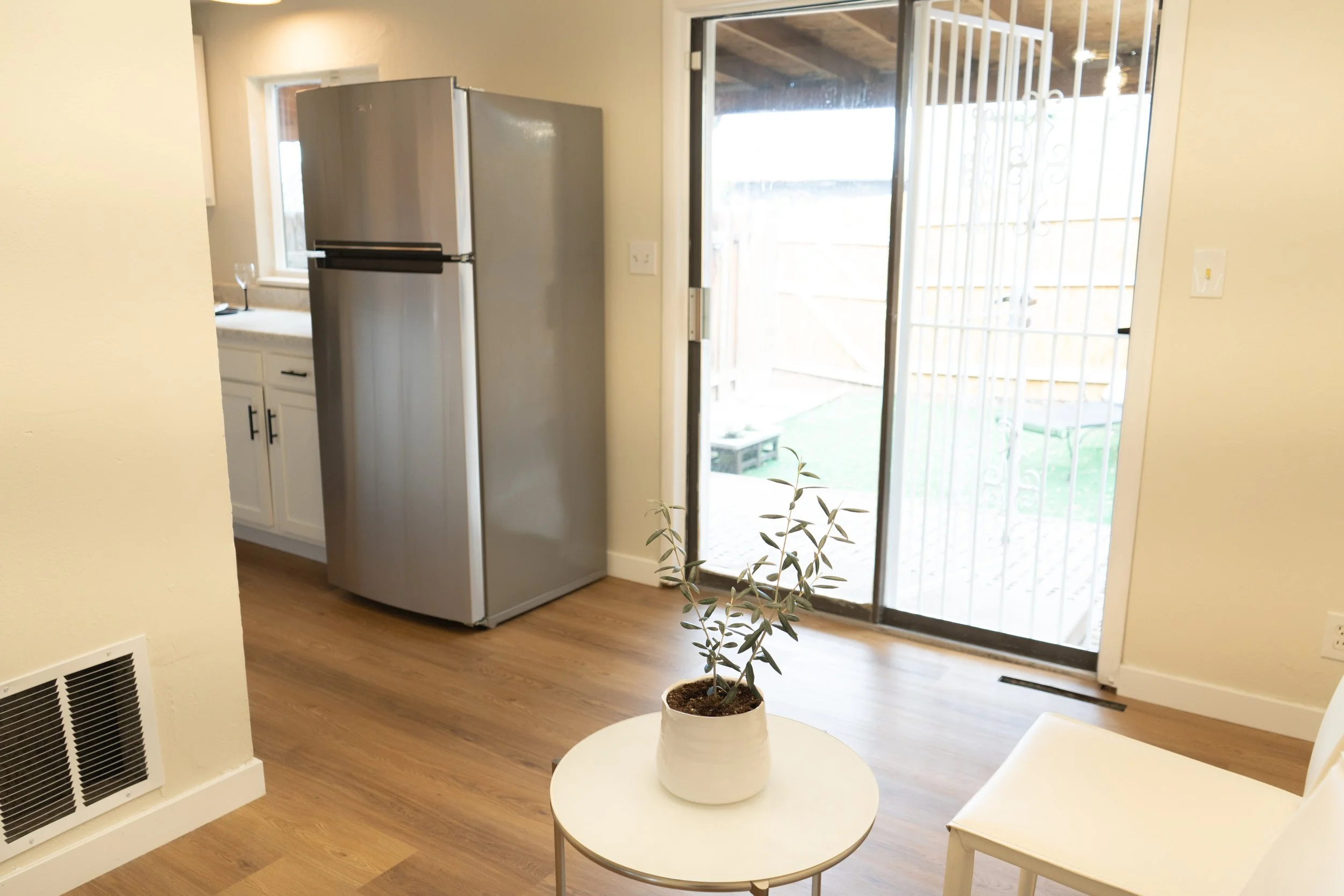 Kitchen corner with a stainless steel refrigerator, white cabinets, a round side table with a potted plant, a white chair, and a sliding glass door leading outside.