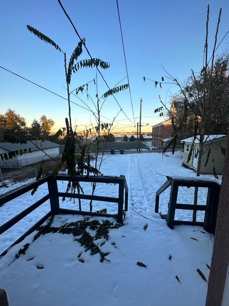 Snow-covered backyard with steps and a broken railing, leafless trees, and clothes hanging on a line, under a clear blue sky at sunset.