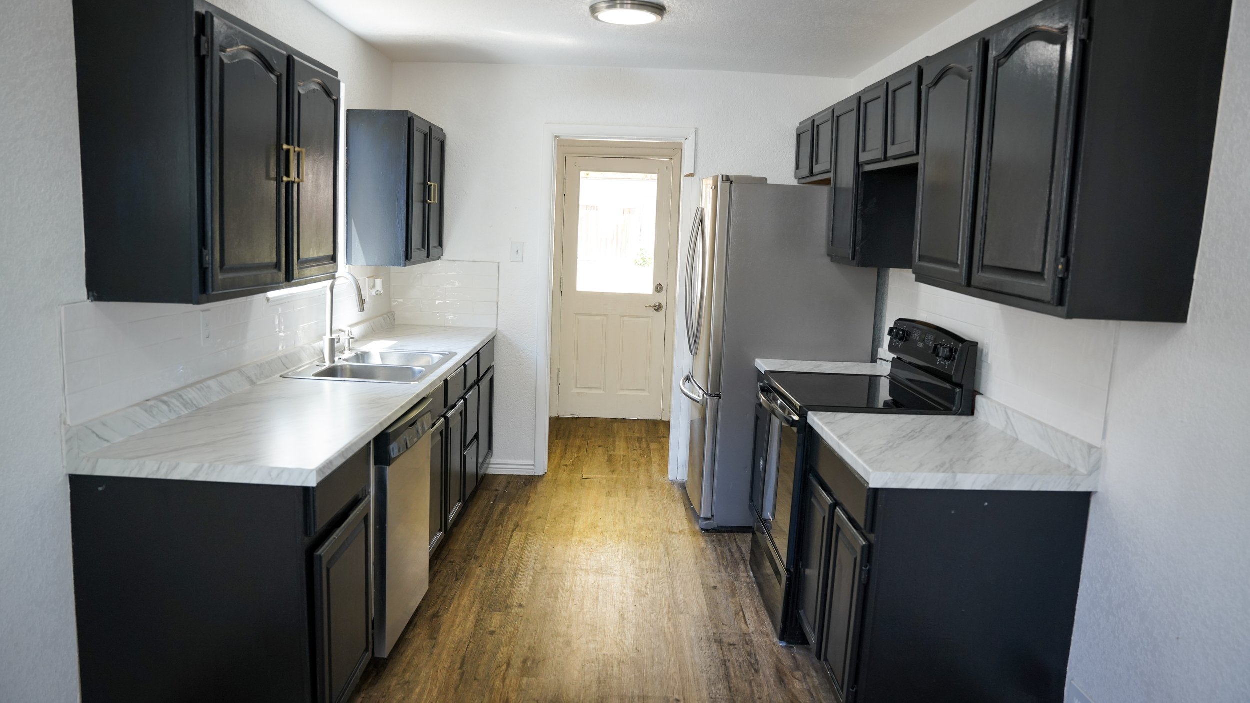 Kitchen with black cabinets, white countertop, stainless steel appliances, and a wooden floor, view of back door.