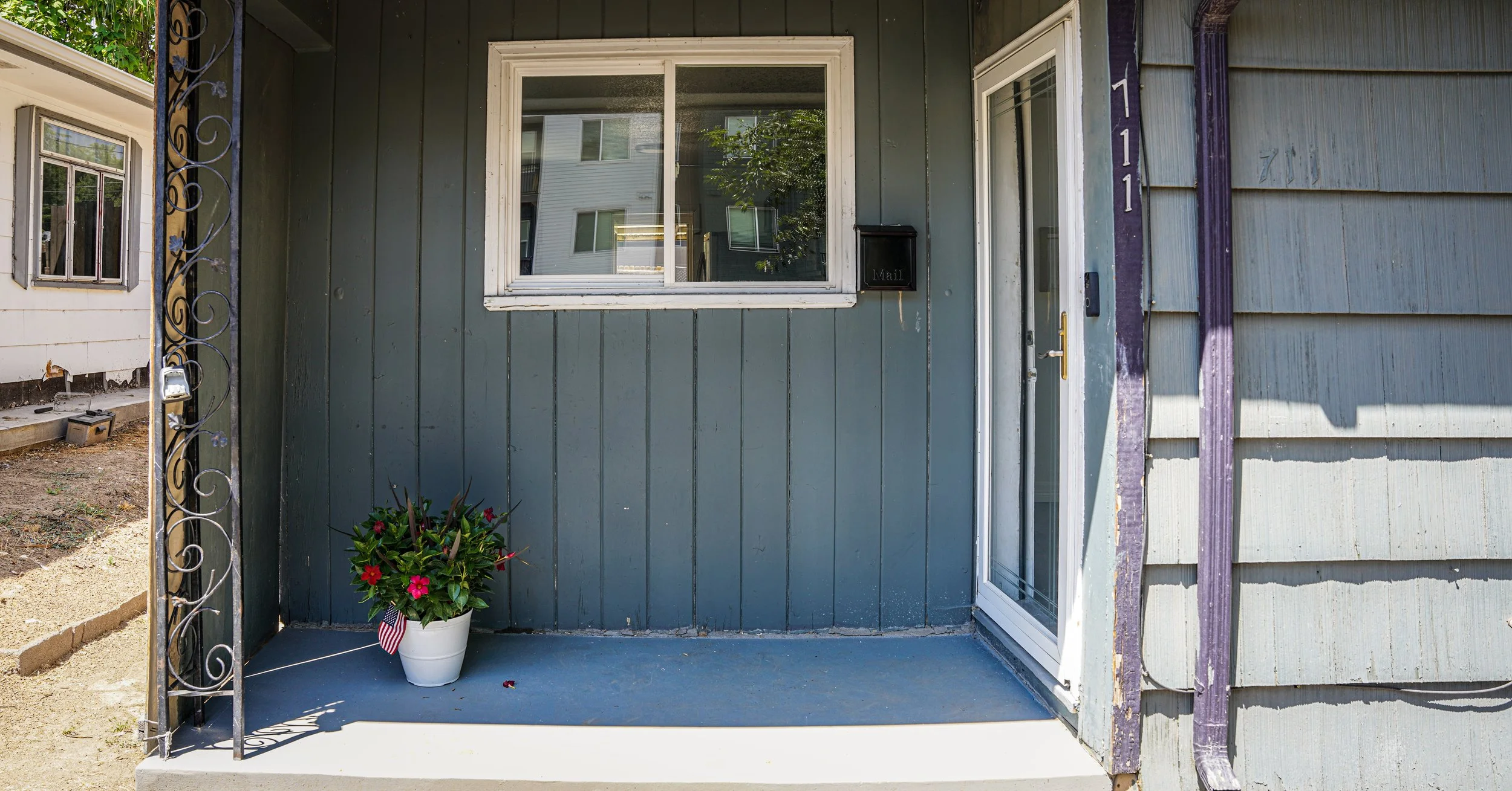 A small front porch with a gray painted wall, a window, a black mailbox labeled 'Mail', a white door, a flower pot with pink flowers and a small American flag, and a decorative metal railing on the left side.