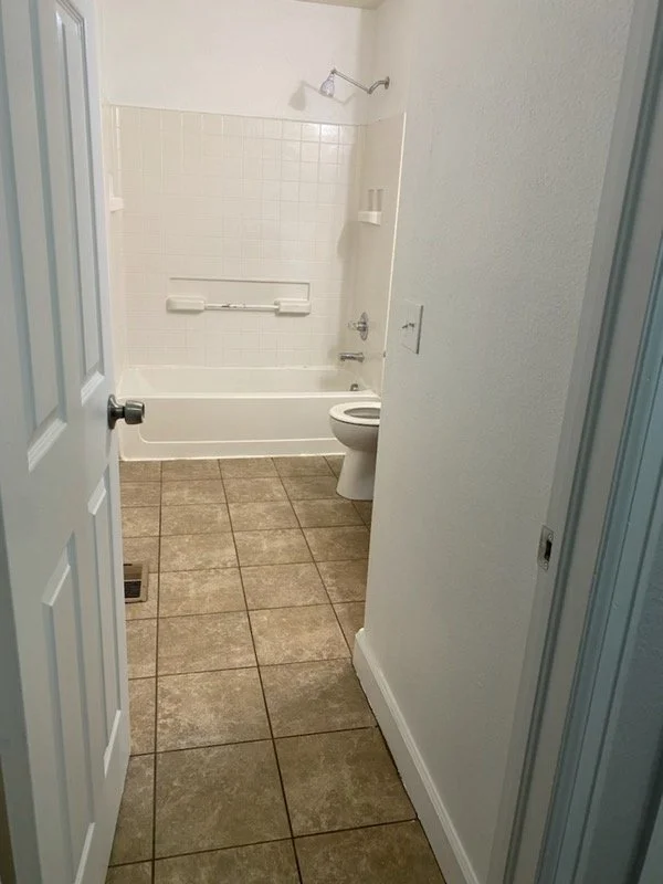 Bathroom with beige tiled floor, white bathtub with grab bar, white walls, and a toilet next to the bathtub, shown through an open door.