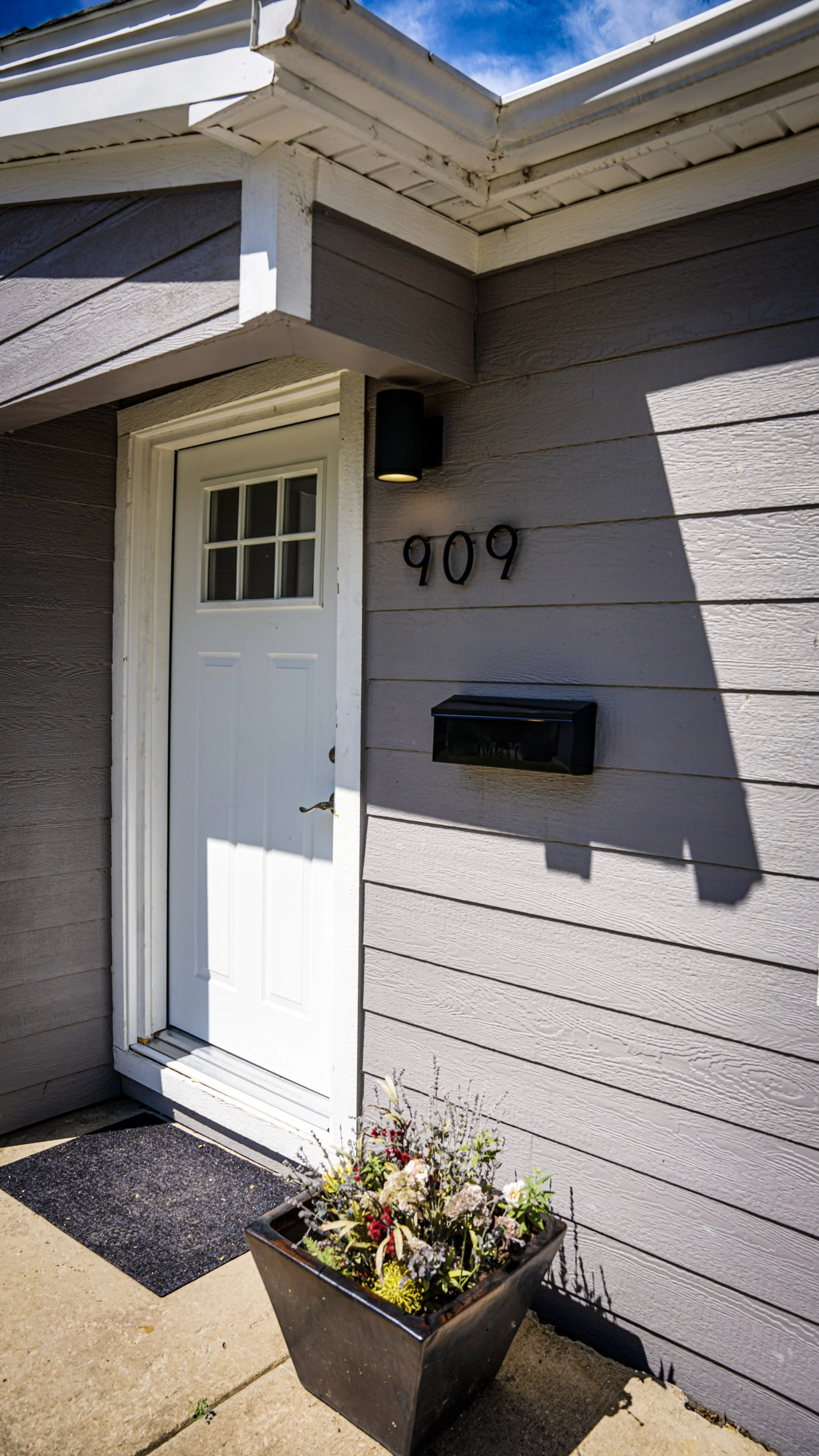 Exterior of a house with gray horizontal siding, a white door with small window panes, black house number '909' mounted on the wall, black mailbox, and a planter with flowers on the concrete porch.
