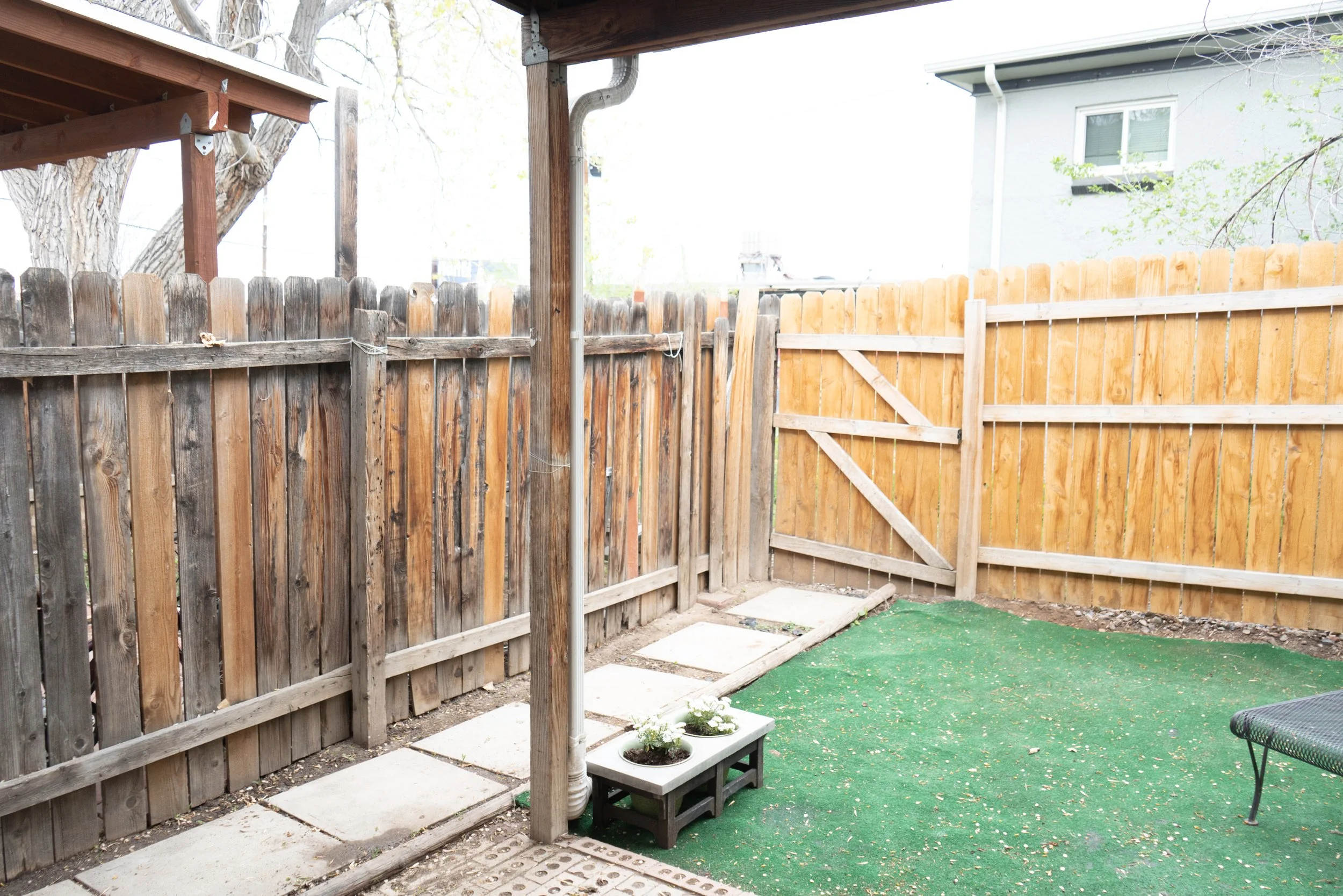 Small fenced backyard with a mix of weathered and new wooden fencing, green artificial grass, potted plants, and outdoor furniture.
