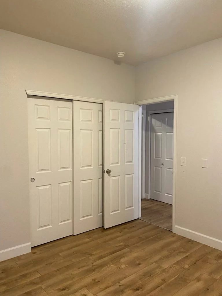 Empty room with white walls, hardwood floors, closet with sliding doors, and a door leading to another room.