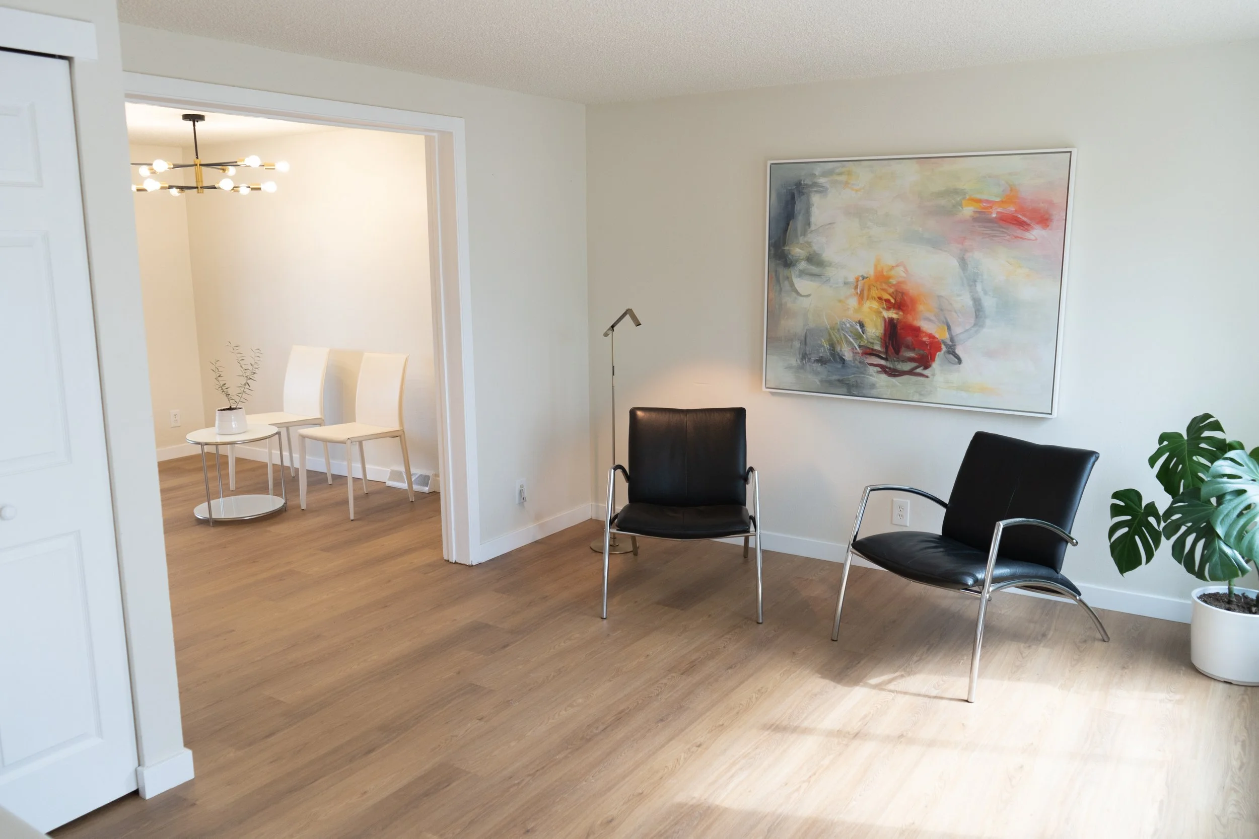 Living room with two black chairs, a potted plant, and abstract painting on the wall. Adjacent dining area with two white chairs and a round side table.