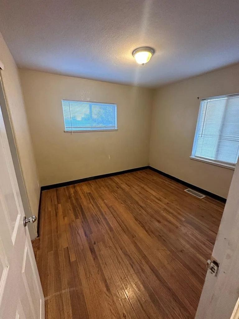 Empty room with hardwood floors, two windows with blinds, and a ceiling light fixture.