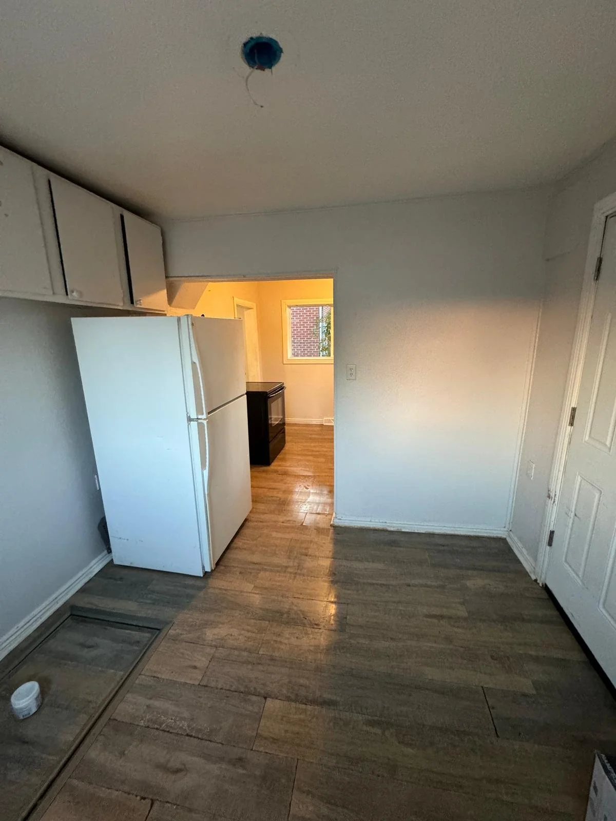 Empty kitchen with white refrigerator, wooden floor, white cabinets, doorway leading to another room with a window, ceiling light fixture missing cover.