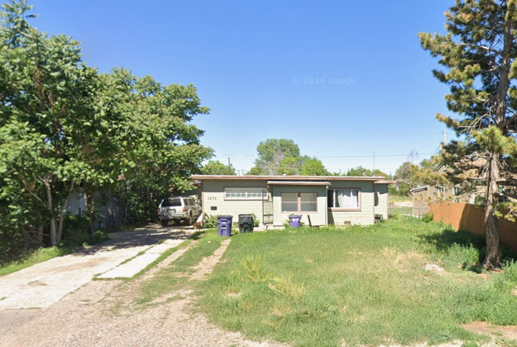 Front yard of a single-story house with a driveway, parked vehicle, trash bins, trees, and a clear blue sky.