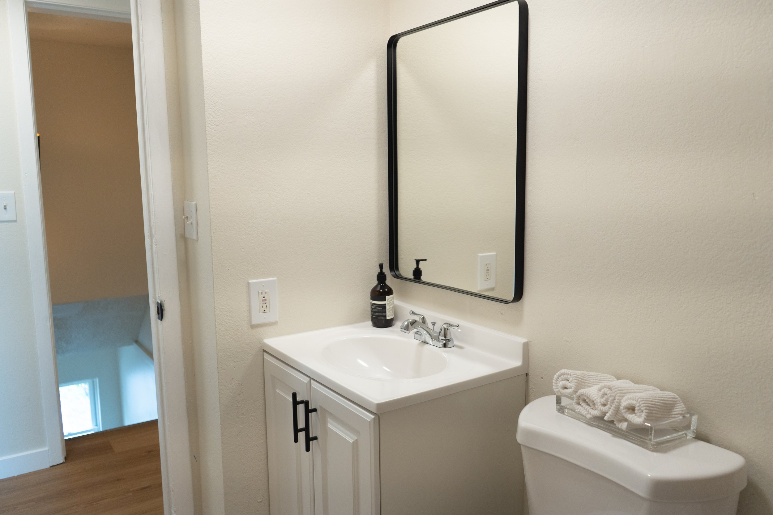 Bathroom with a white vanity, black-framed mirror, soap dispenser, and towel on the toilet tank, with a door leading to stairs and a window visible in the background.