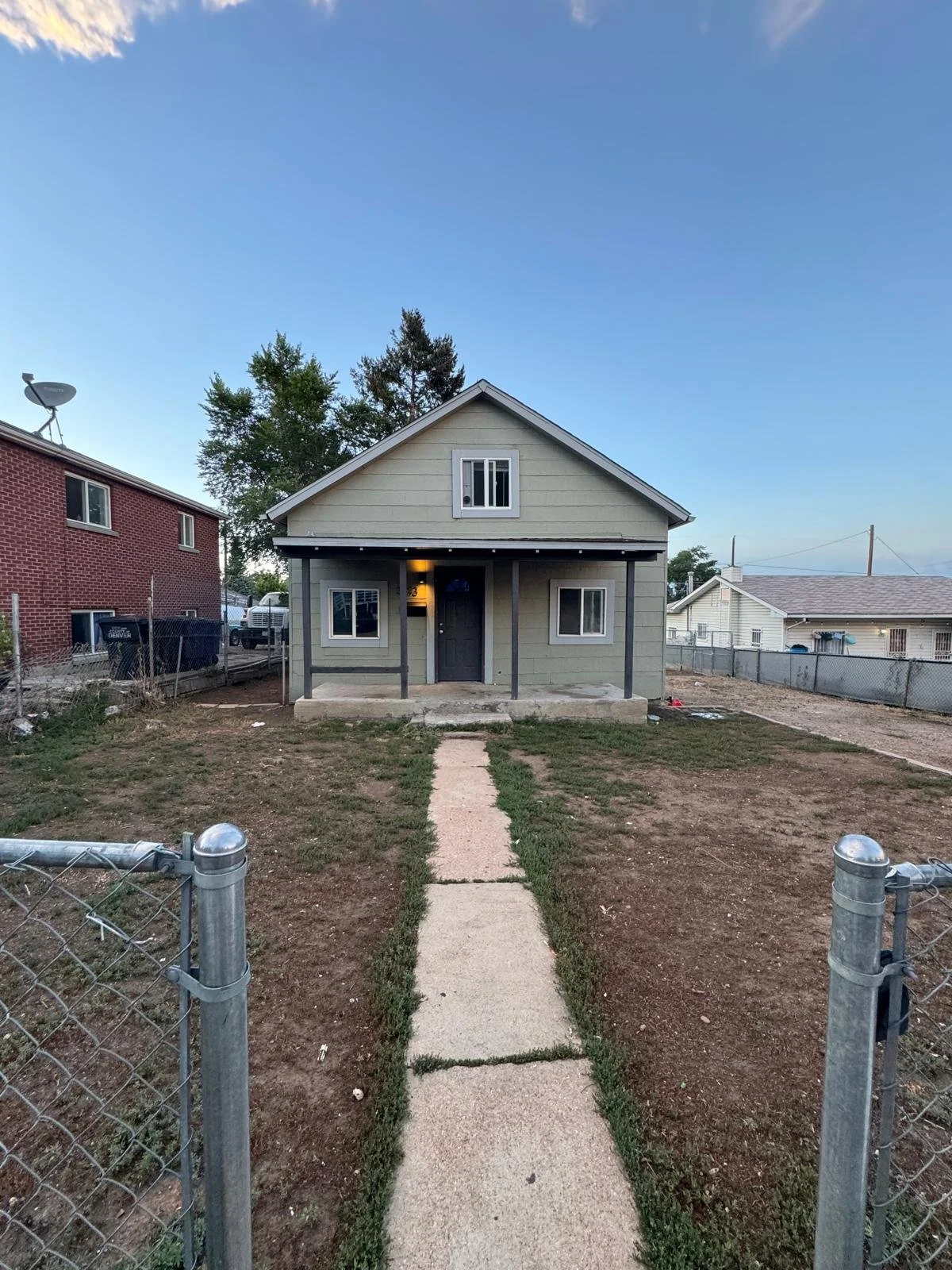 Small house with a porch, two windows in the front, and a single door. The front yard has a concrete walkway and is fenced with a chain-link fence.