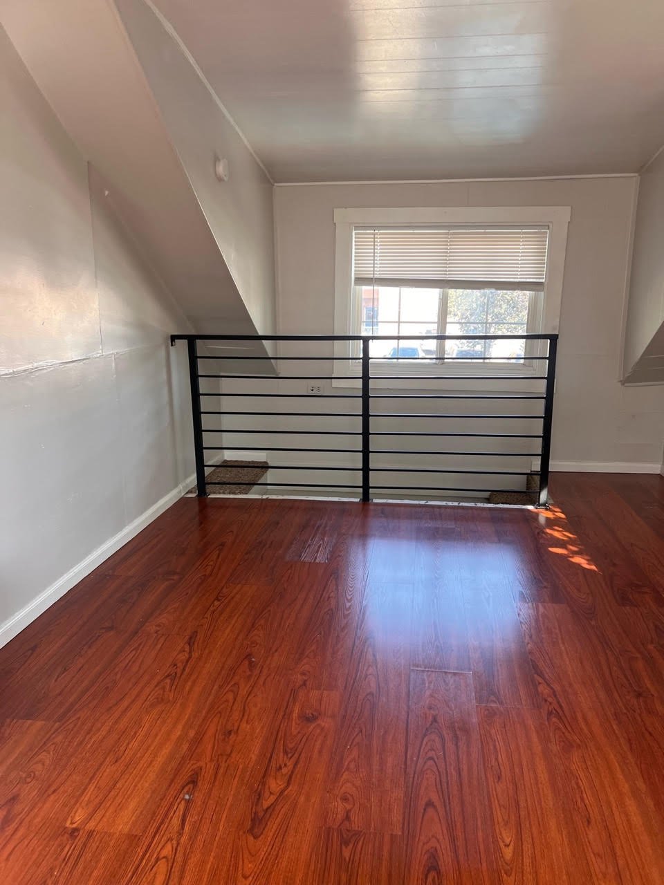 Empty room with wooden floors, white walls, a large window with blinds, and a black metal railing.