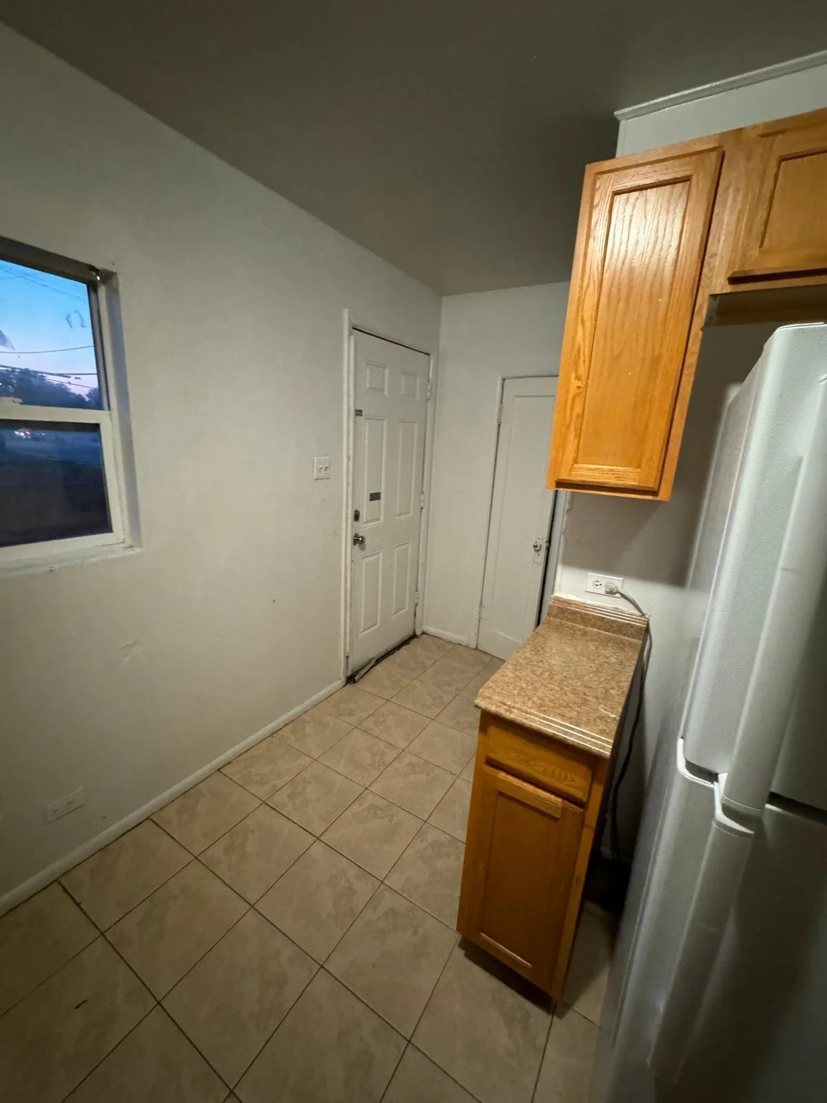 Empty kitchen with a window, tile floor, wooden cabinets, and a white refrigerator.