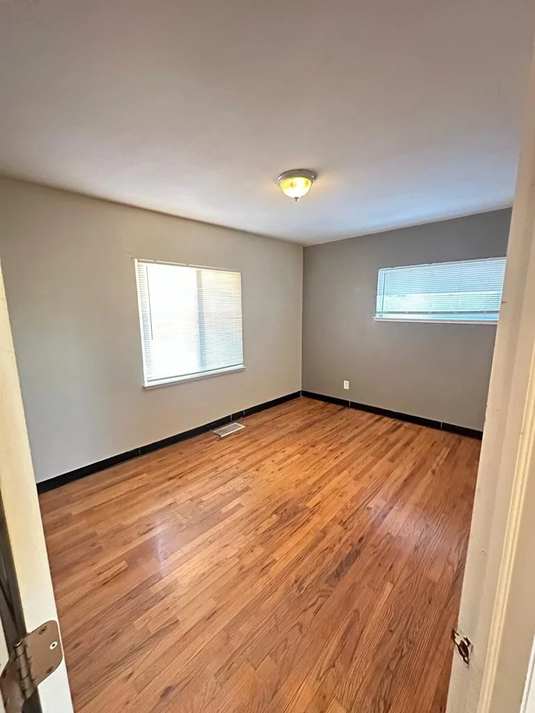 Empty room with hardwood floors, beige walls, two windows with blinds, and a ceiling light fixture.
