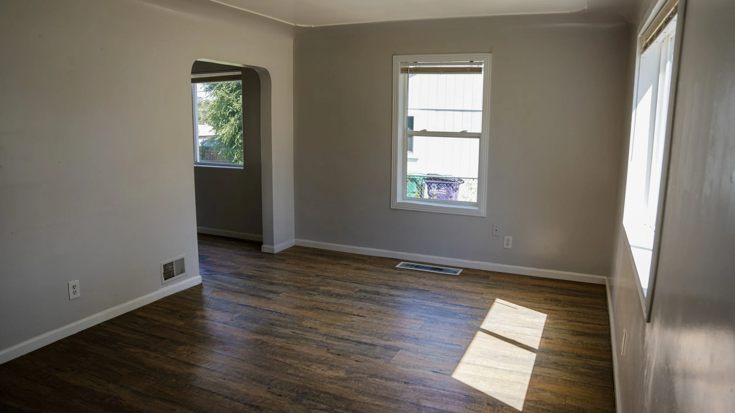 Empty living room with hardwood floors, beige walls, and two windows allowing natural light in.