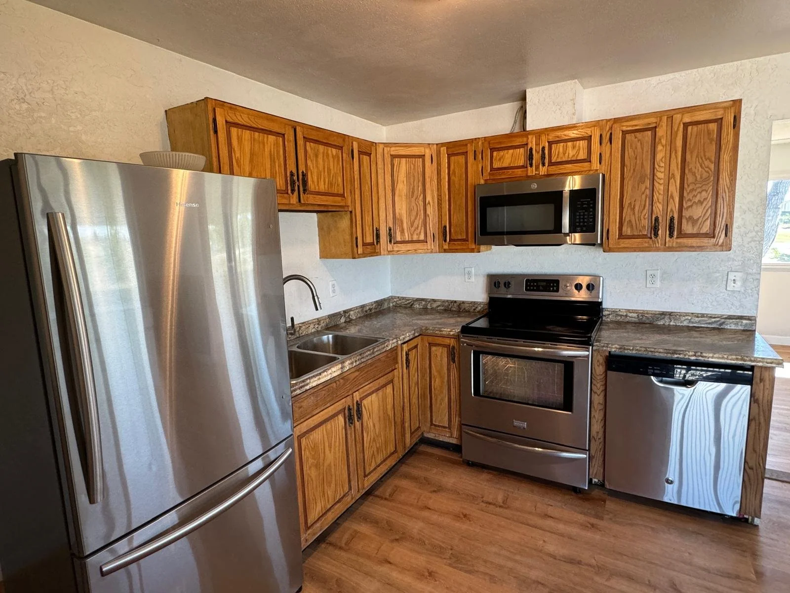 Kitchen with wooden cabinets, stainless steel refrigerator, stove, microwave, and dishwasher, granite countertops, and wood flooring.