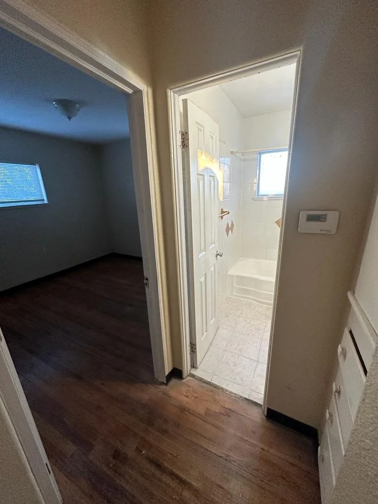 View of a small bathroom with a bathtub and window, seen through an open door from a hallway with wooden flooring.