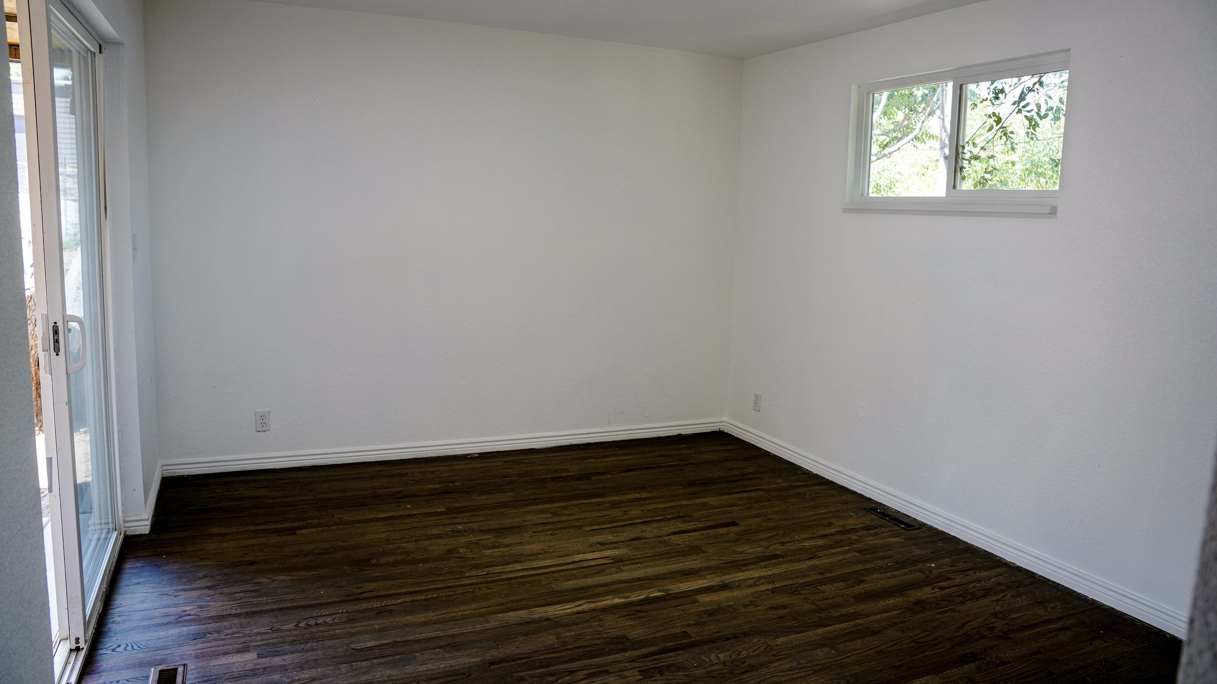 Empty room with white walls, dark wood flooring, a sliding glass door on the left and a window on the right wall showing green trees outside.