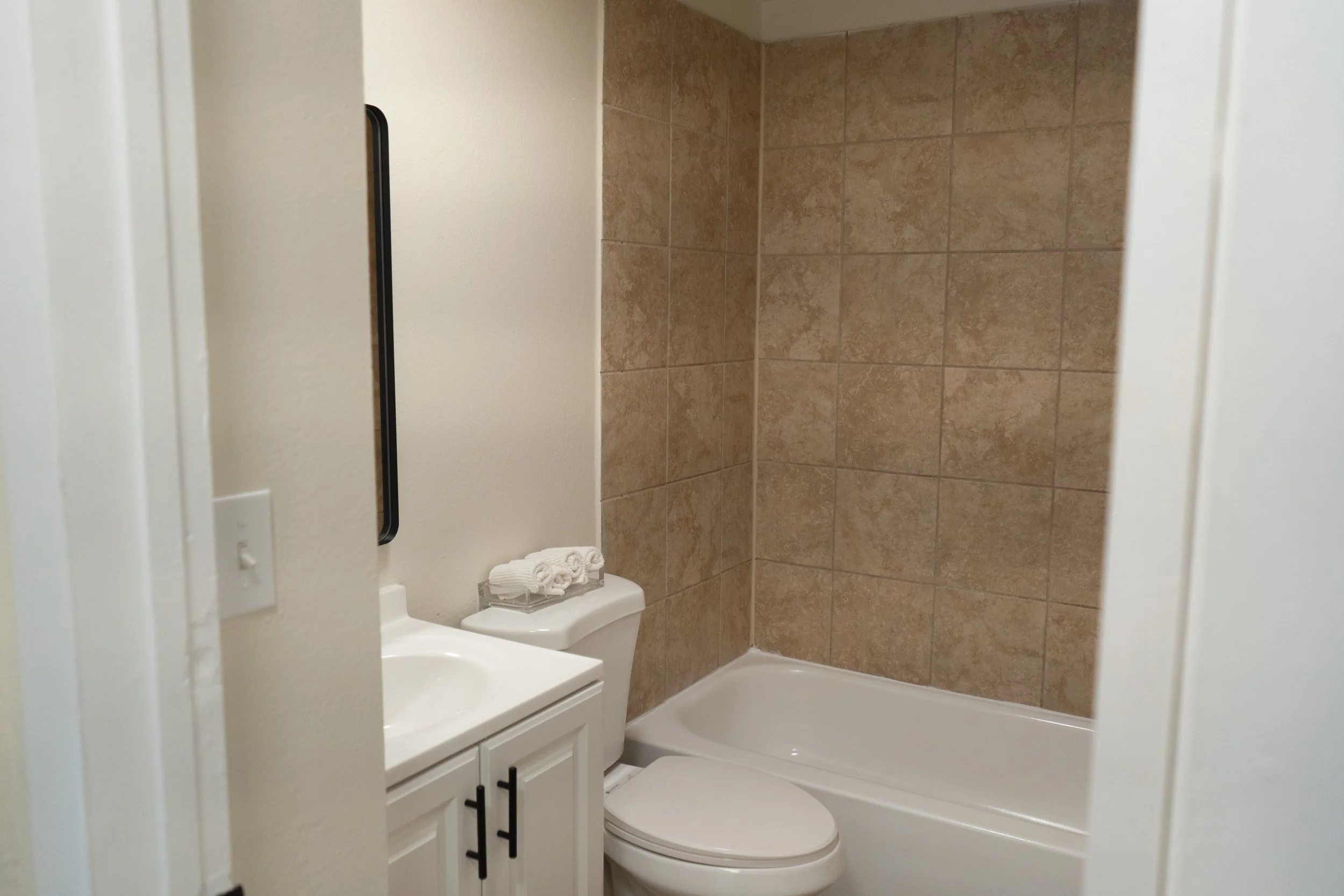 Small bathroom with a bathtub, toilet, and a vanity with a sink, set against a beige tiled wall.